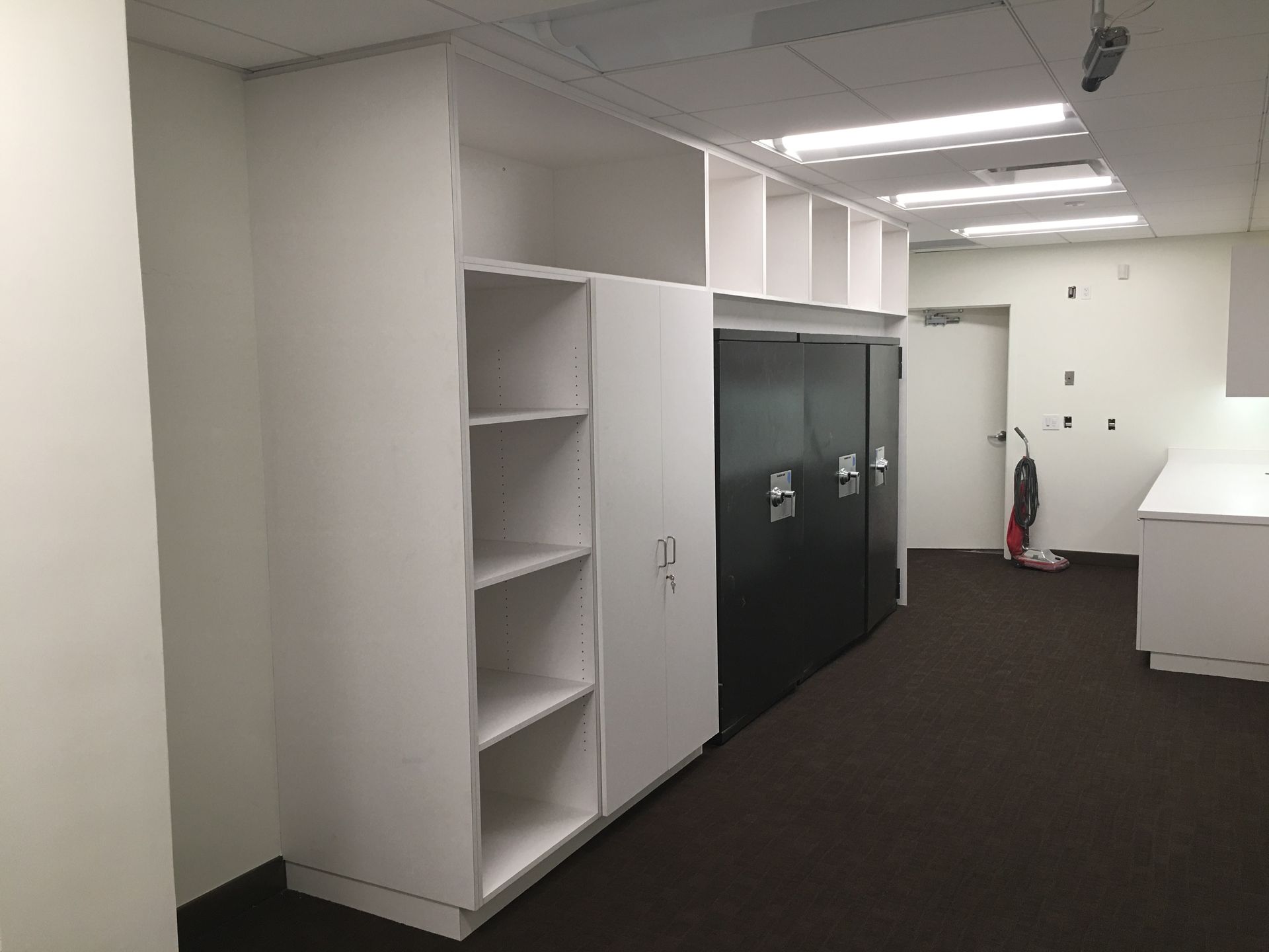 Office interior with white storage cabinets, open shelves, and black lockers. Brown carpet.