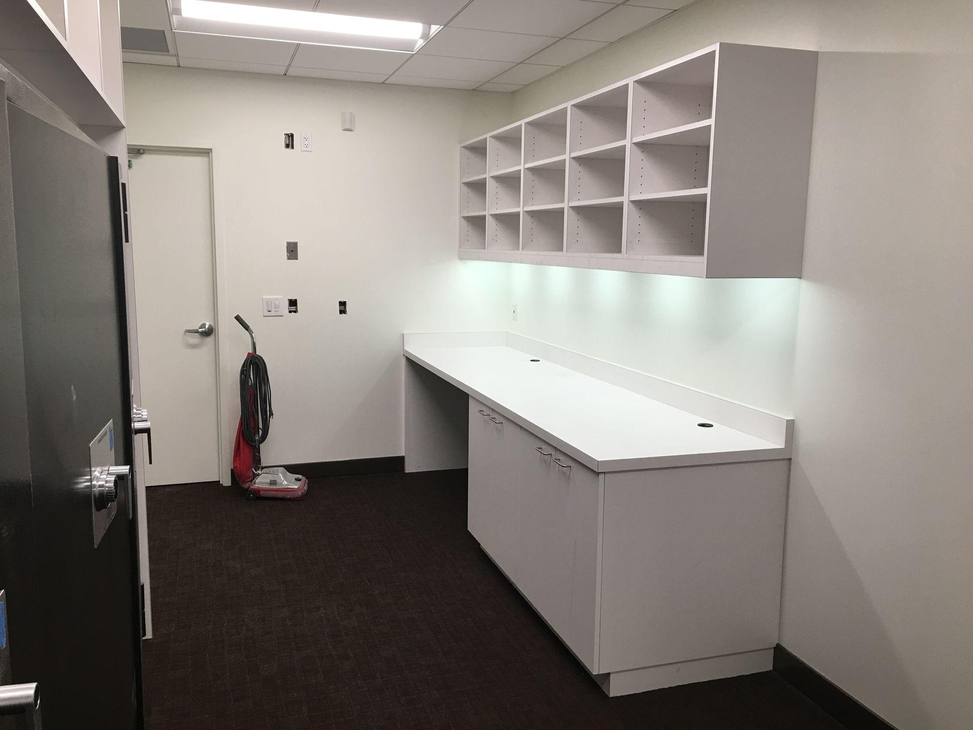 Empty office with white cabinets, shelves, and countertop. Dark carpet and a vacuum cleaner.