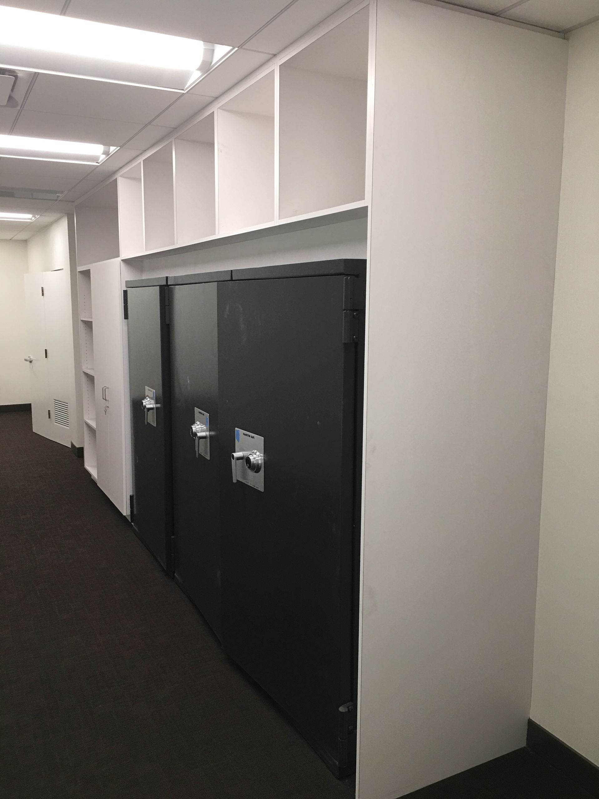 Dark metal lockers built into a wall with white shelving above in a hallway.