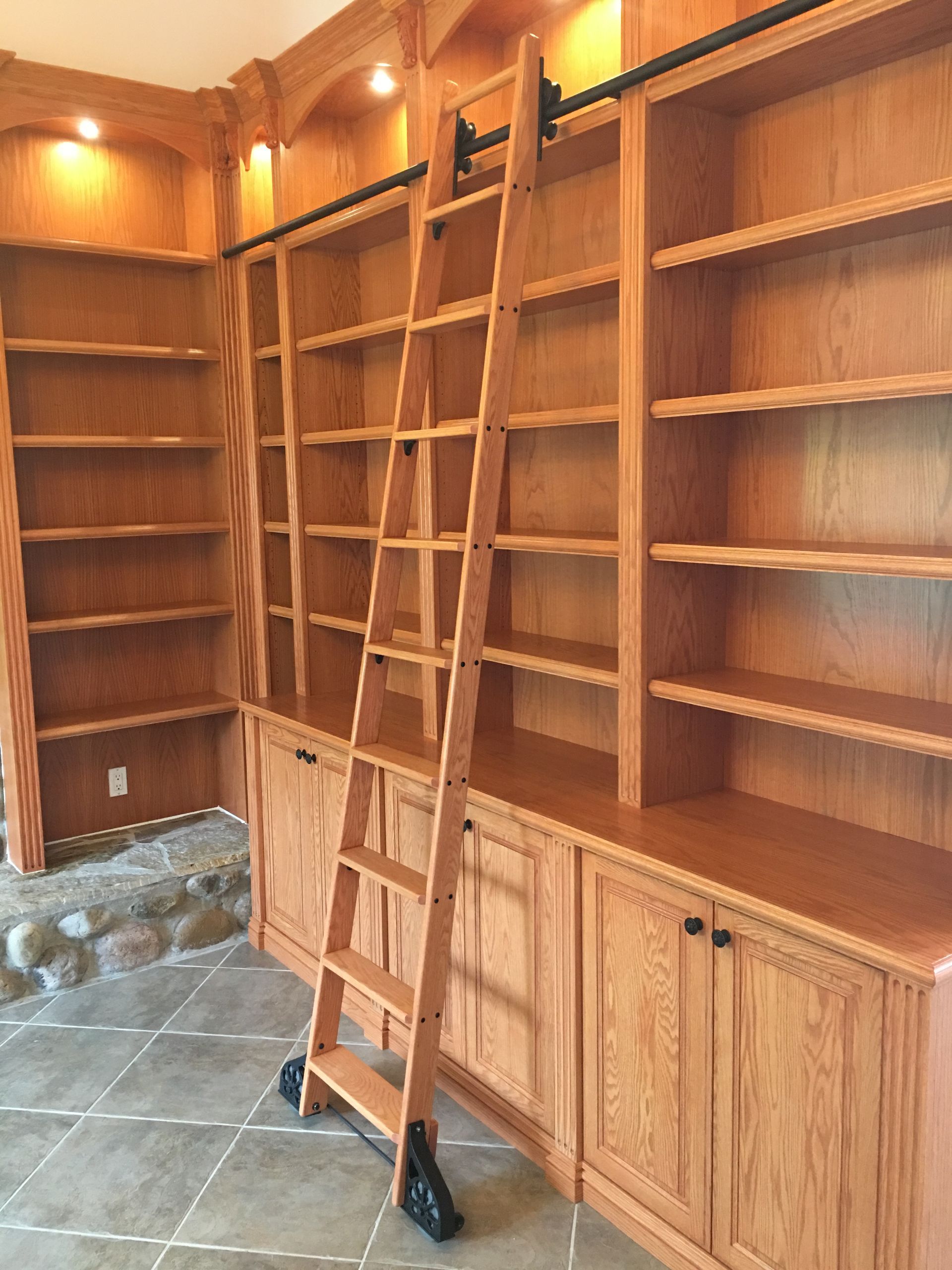 Wooden library with rolling ladder. Bookshelves with storage cabinets. Brown wood, black hardware.