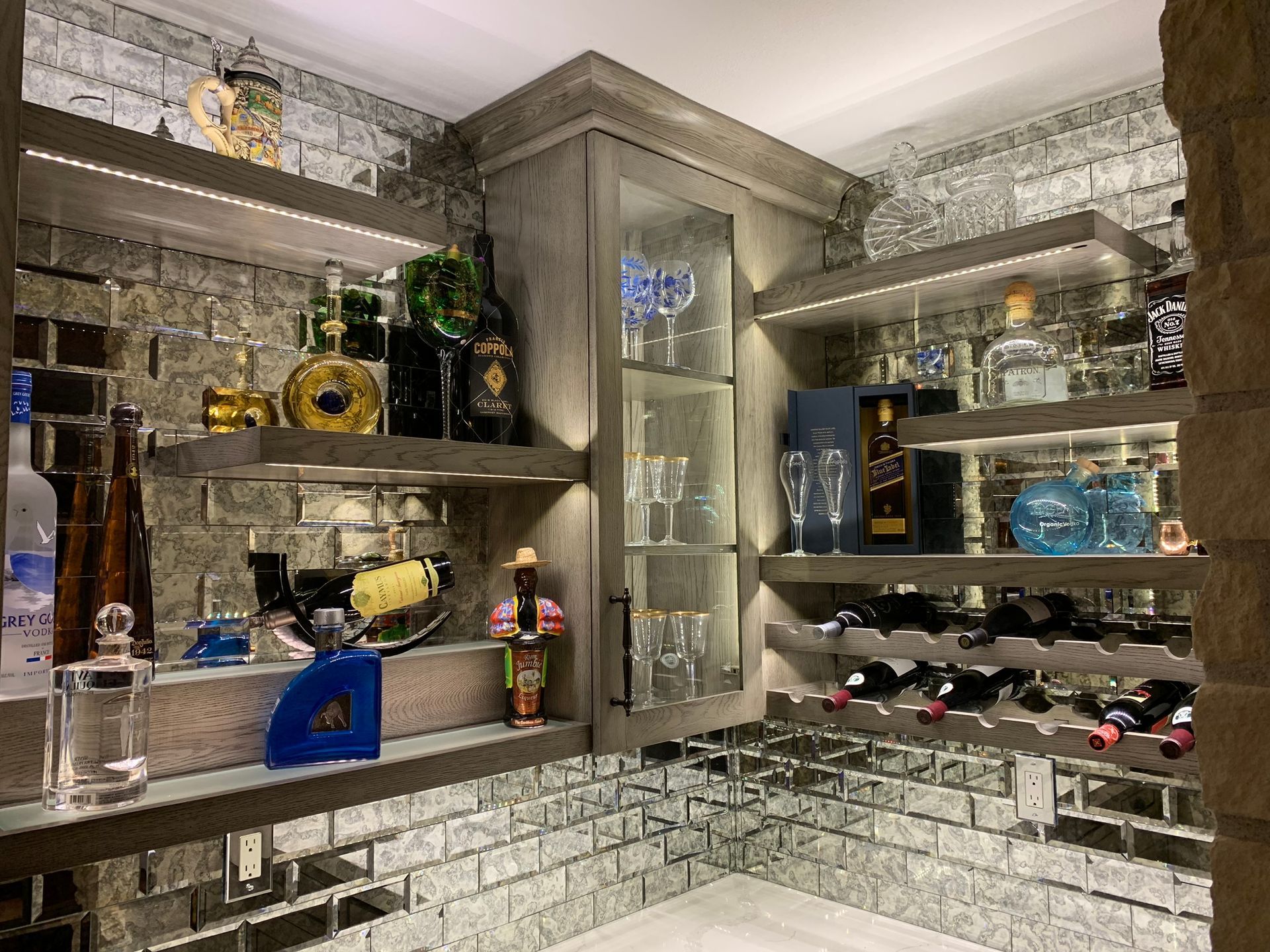 A home bar with mirrored brick walls, wooden shelves, and liquor bottles. Built-in cabinet holds glassware.
