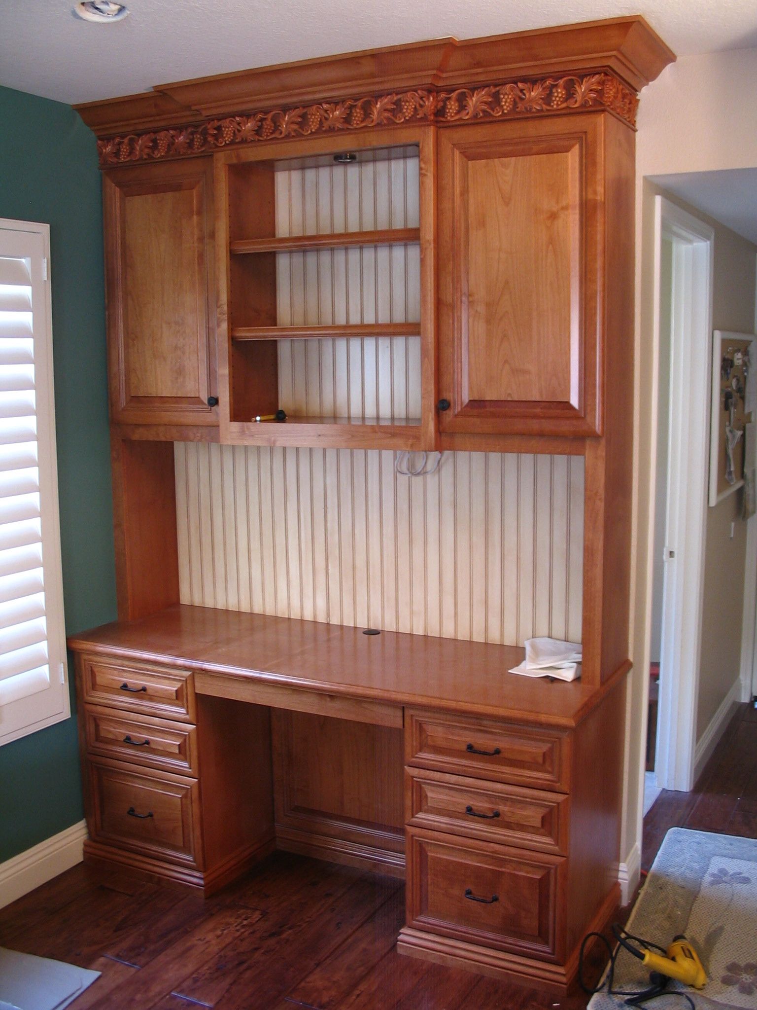 Wooden desk with upper cabinets and carved trim, built-in against a wall with beadboard and green paint.