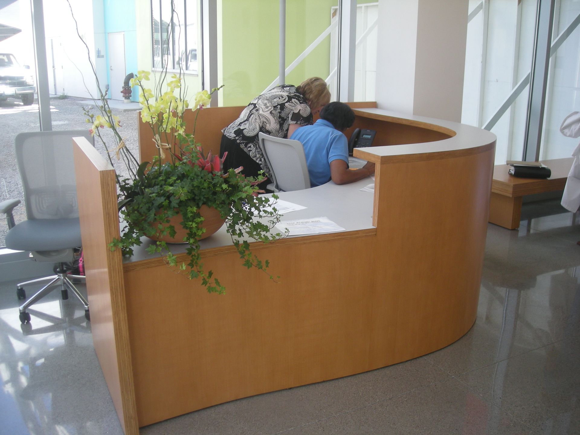 Reception desk with two people interacting; flowers in a pot, light wood finish.
