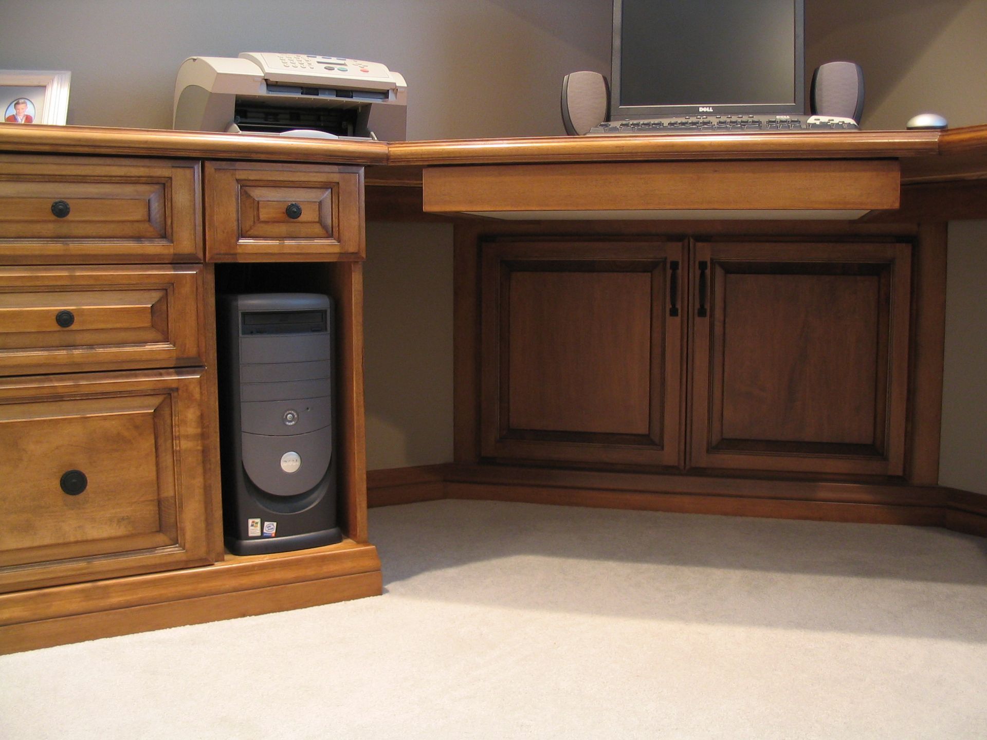 Wooden desk with built-in computer tower and printer. Cabinets with closed doors. Beige carpet.