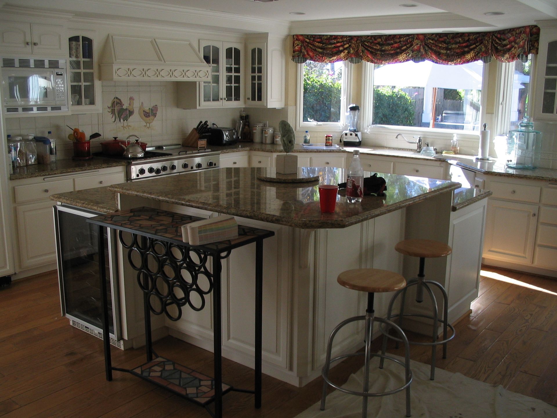 Cream-colored kitchen with granite countertops, island with bar stools, window with valance, and built-in appliances.