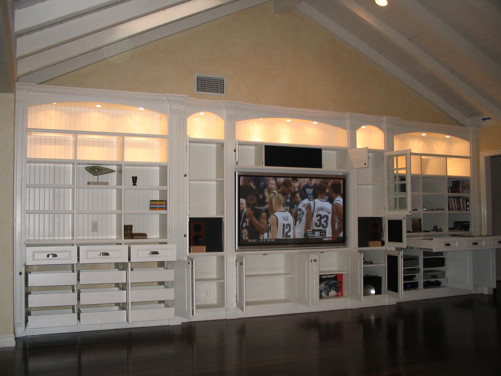 Built-in white entertainment center with a TV, shelving, drawers, and overhead lighting in a room with a vaulted ceiling.