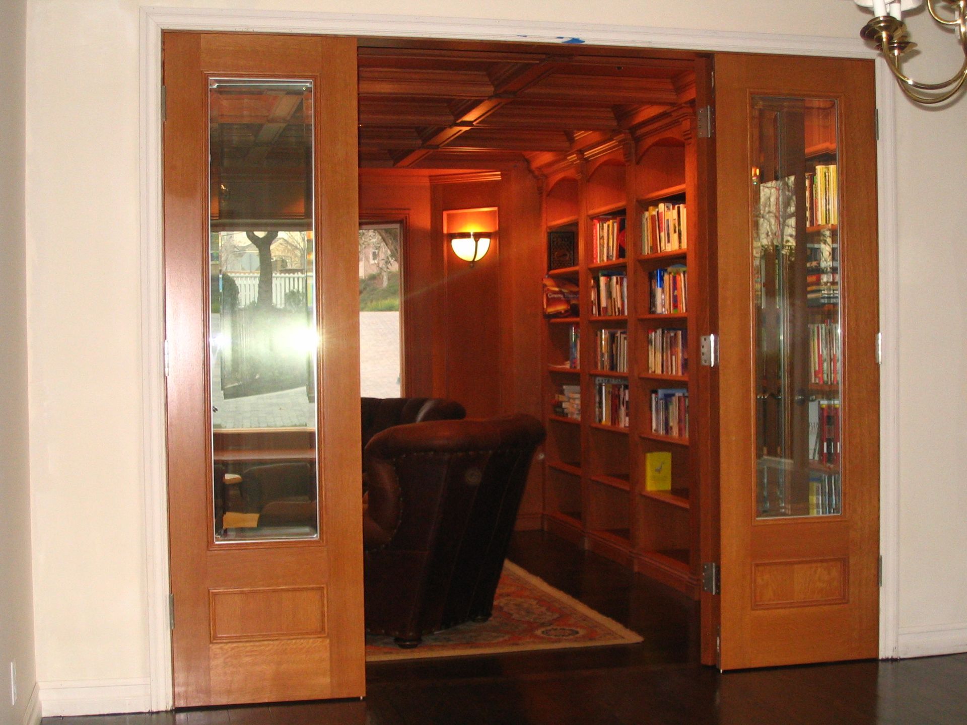 Wooden double doors open to a cozy library with built-in bookshelves, dark furniture, and warm lighting.