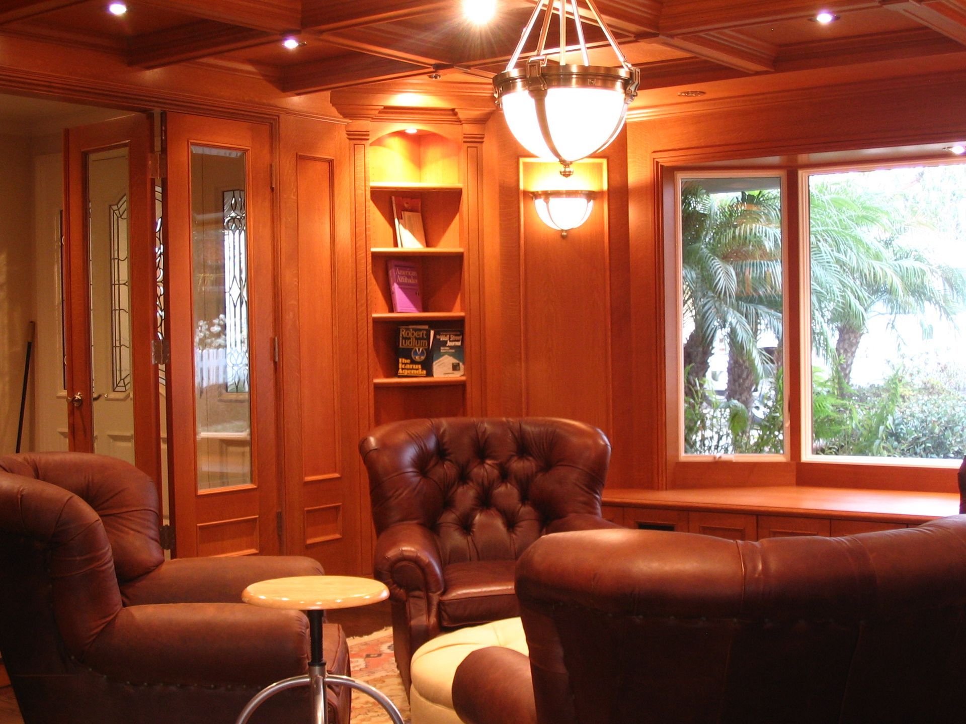 Cozy wood-paneled room with leather armchairs, a small table, a built-in bookshelf, and a window overlooking greenery.