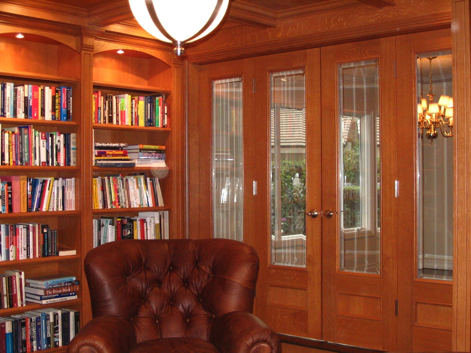 Library with built-in bookshelves, leather armchair, French doors with glass panes. Warm wood tones.