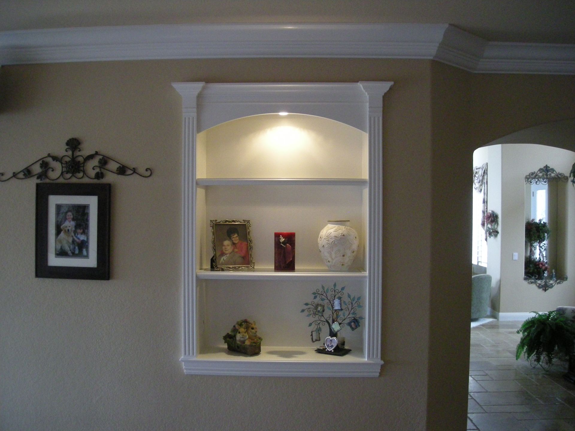 Built-in white shelf with arched top, lit with a light. Displays decorative items on shelves, set in beige wall.