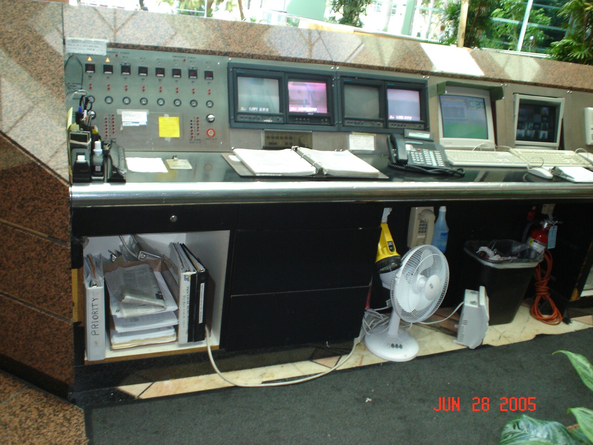 Control panel with monitors and equipment, with a fan and trash can in front.
