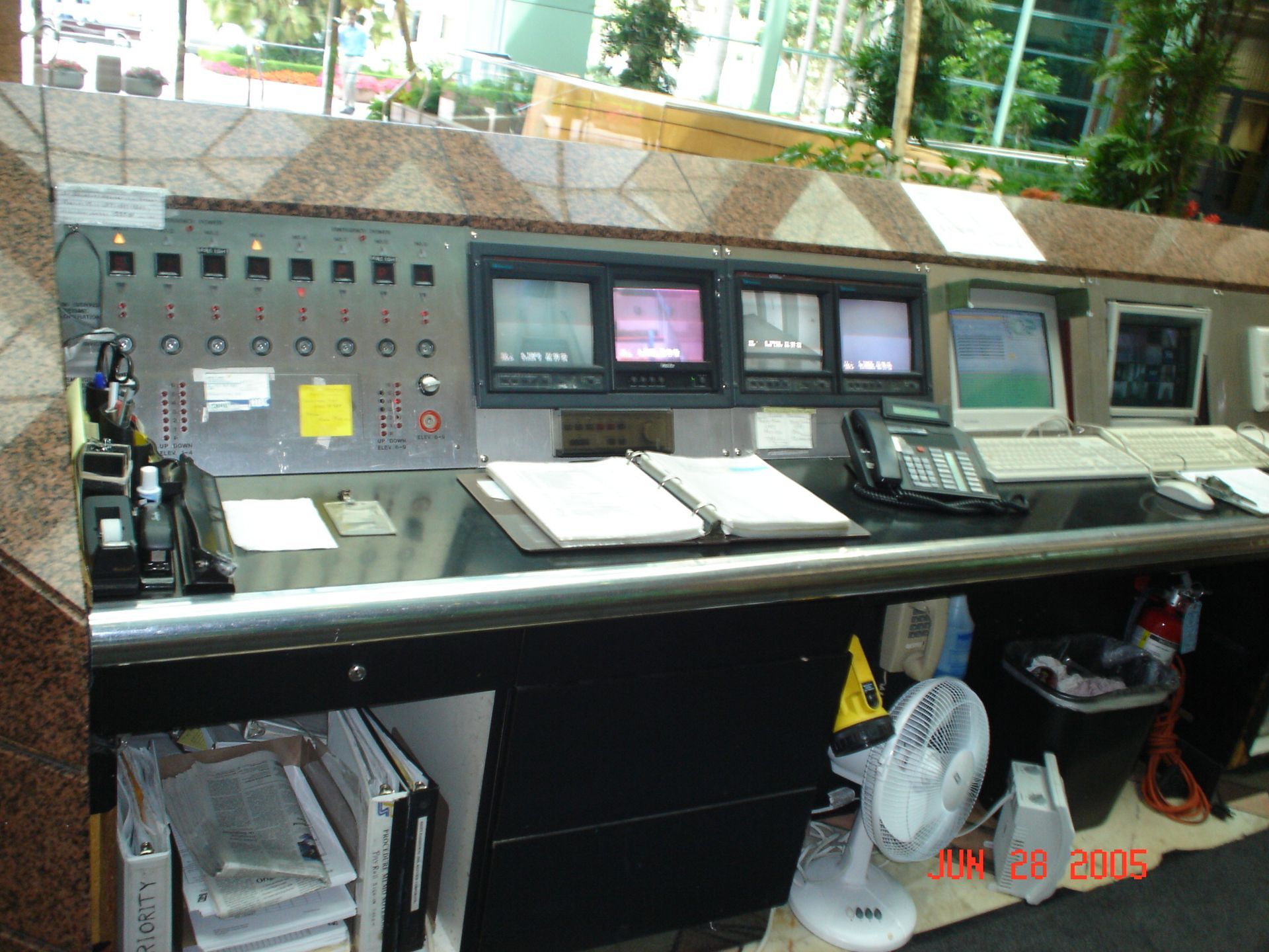 Control panel with screens, buttons, and phones on a desk. White fan, trash can, and folders visible.