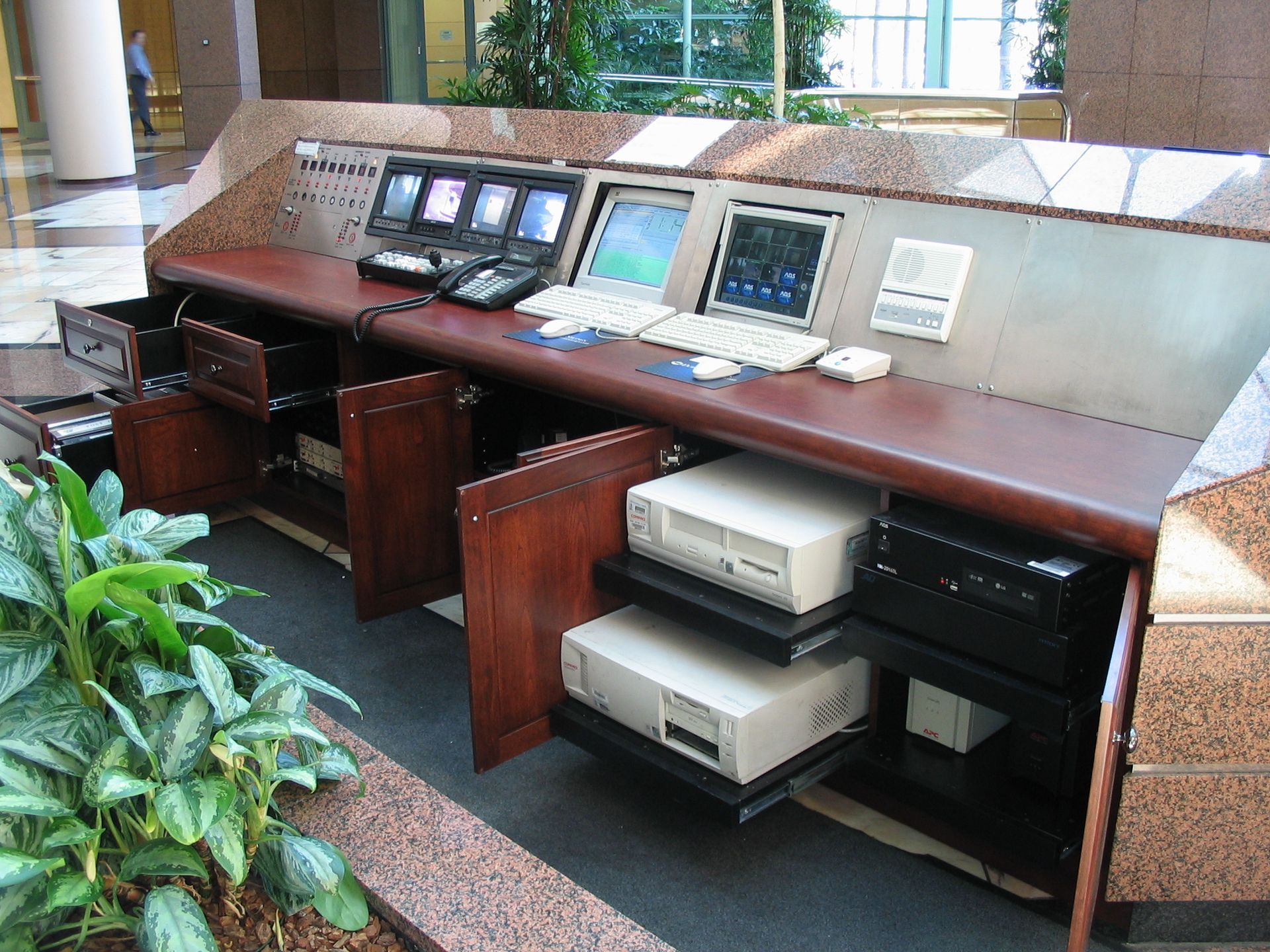 Reception desk with computers, screens, and plants. Brown wood and beige brick.