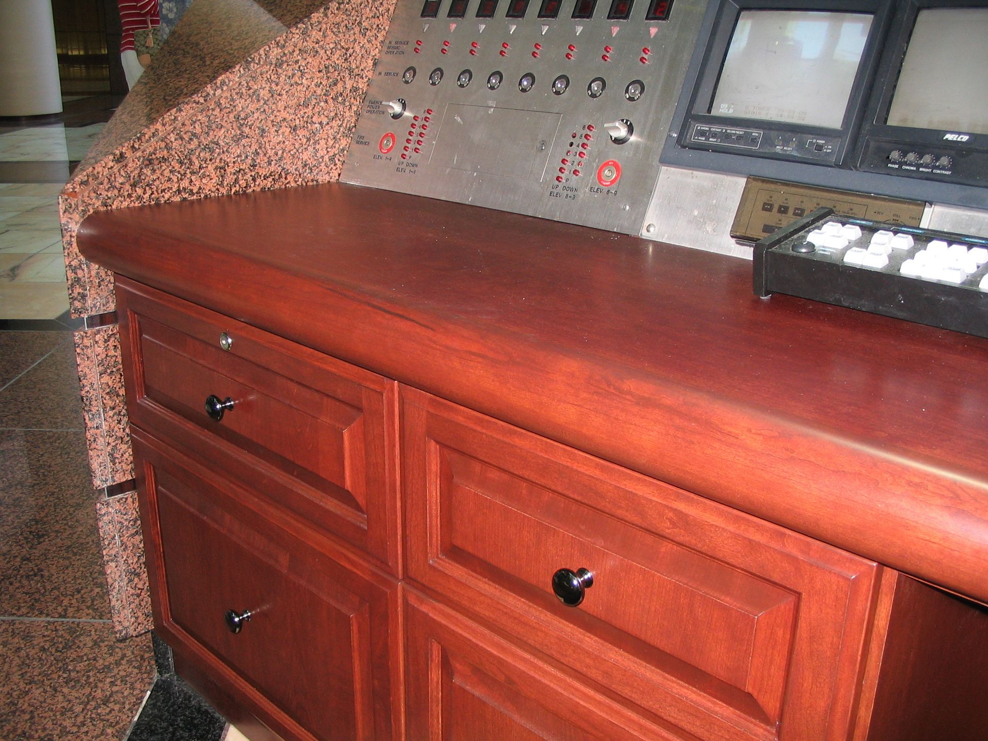 Wooden desk with drawers and knobs, next to control panel and monitors, in an interior space.