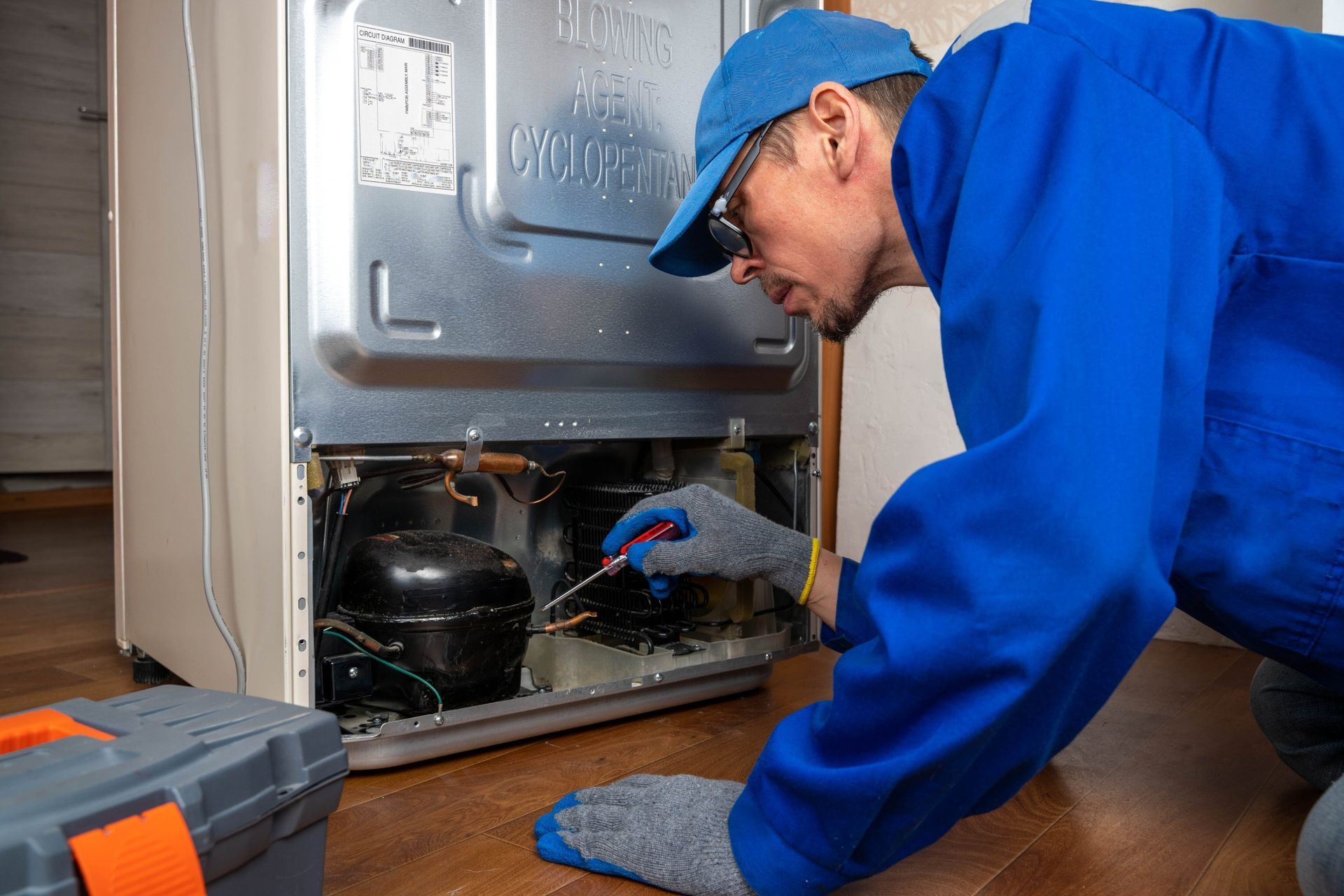 Technician in blue uniform repairing a refrigerator compressor with tools on the floor