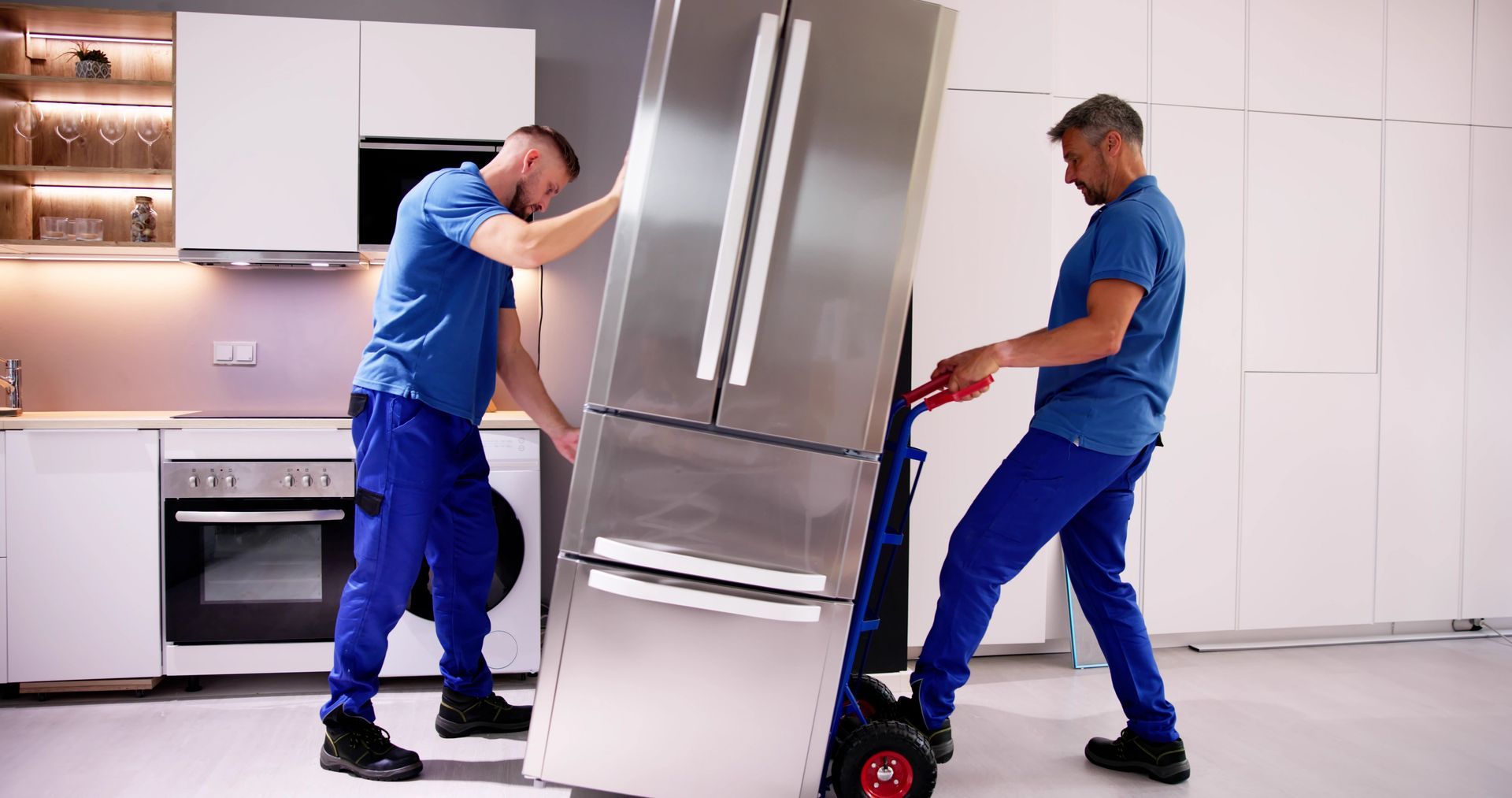 Two movers pushing a stainless steel refrigerator through a modern kitchen on a dolly