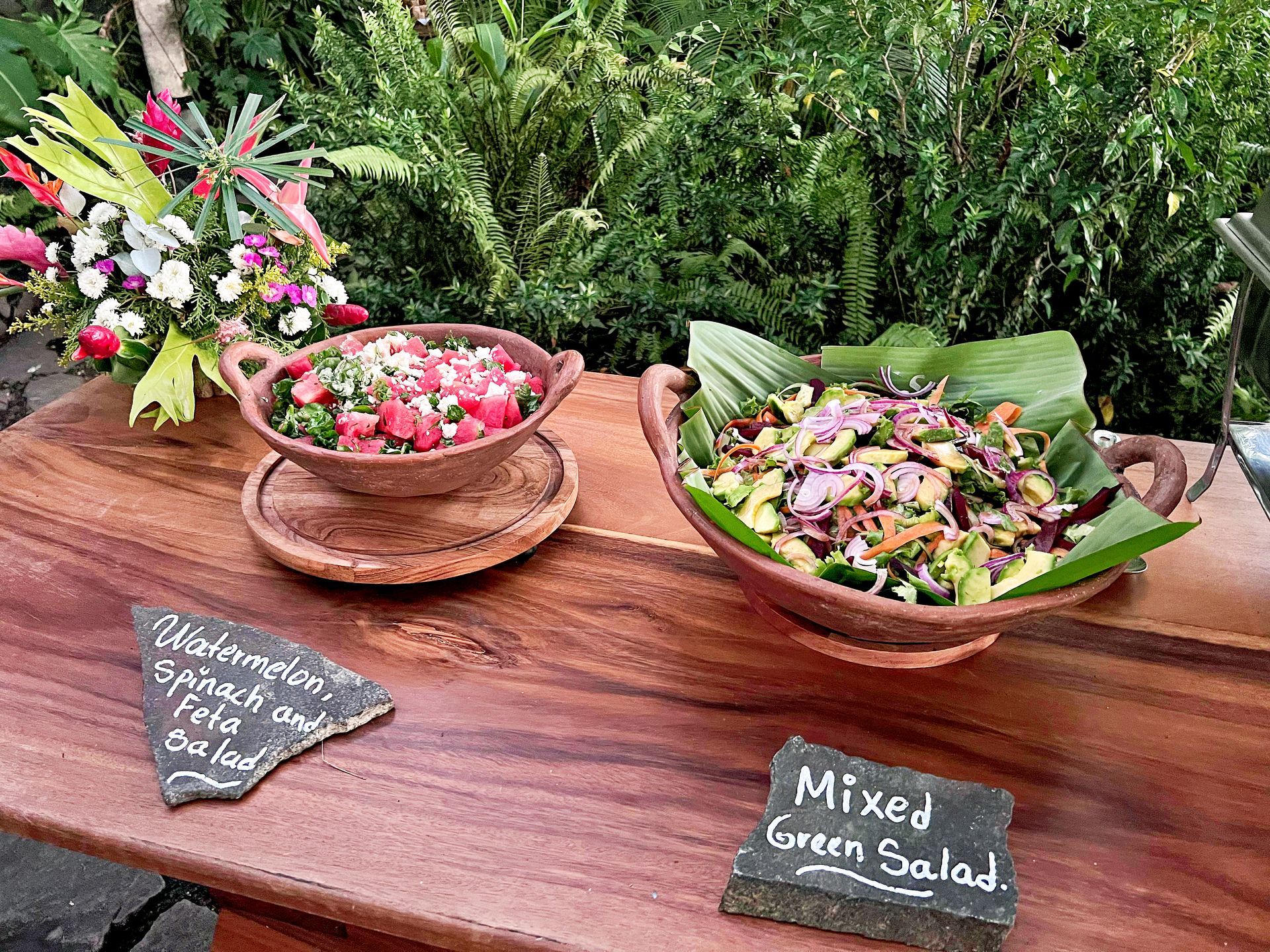 Two salad dishes on a wooden table. One is watermelon and spinach; the other is a mixed green salad.