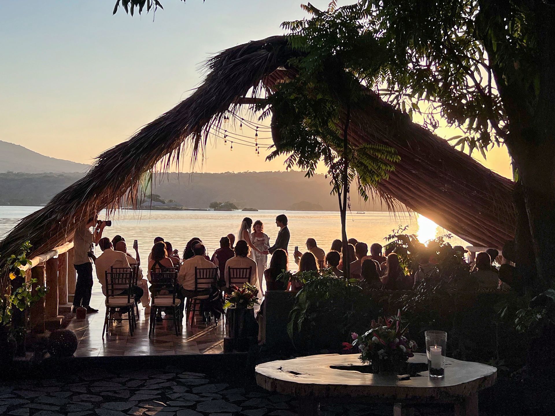 Wedding ceremony at sunset by the water with guests seated; couple under a thatched roof; photographer present.
