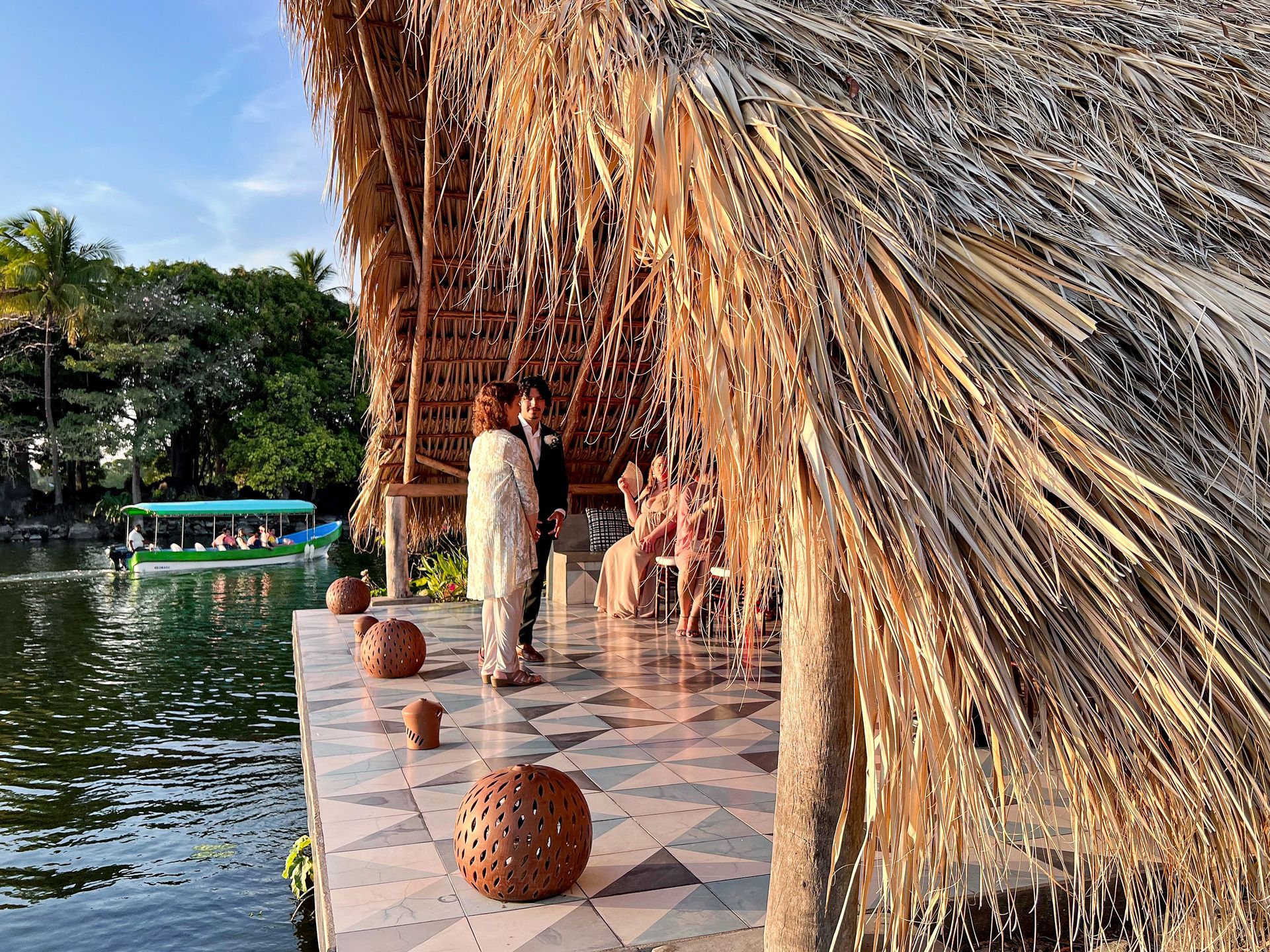 Couple getting married by a lake, under a thatched roof. Attendees seated nearby.