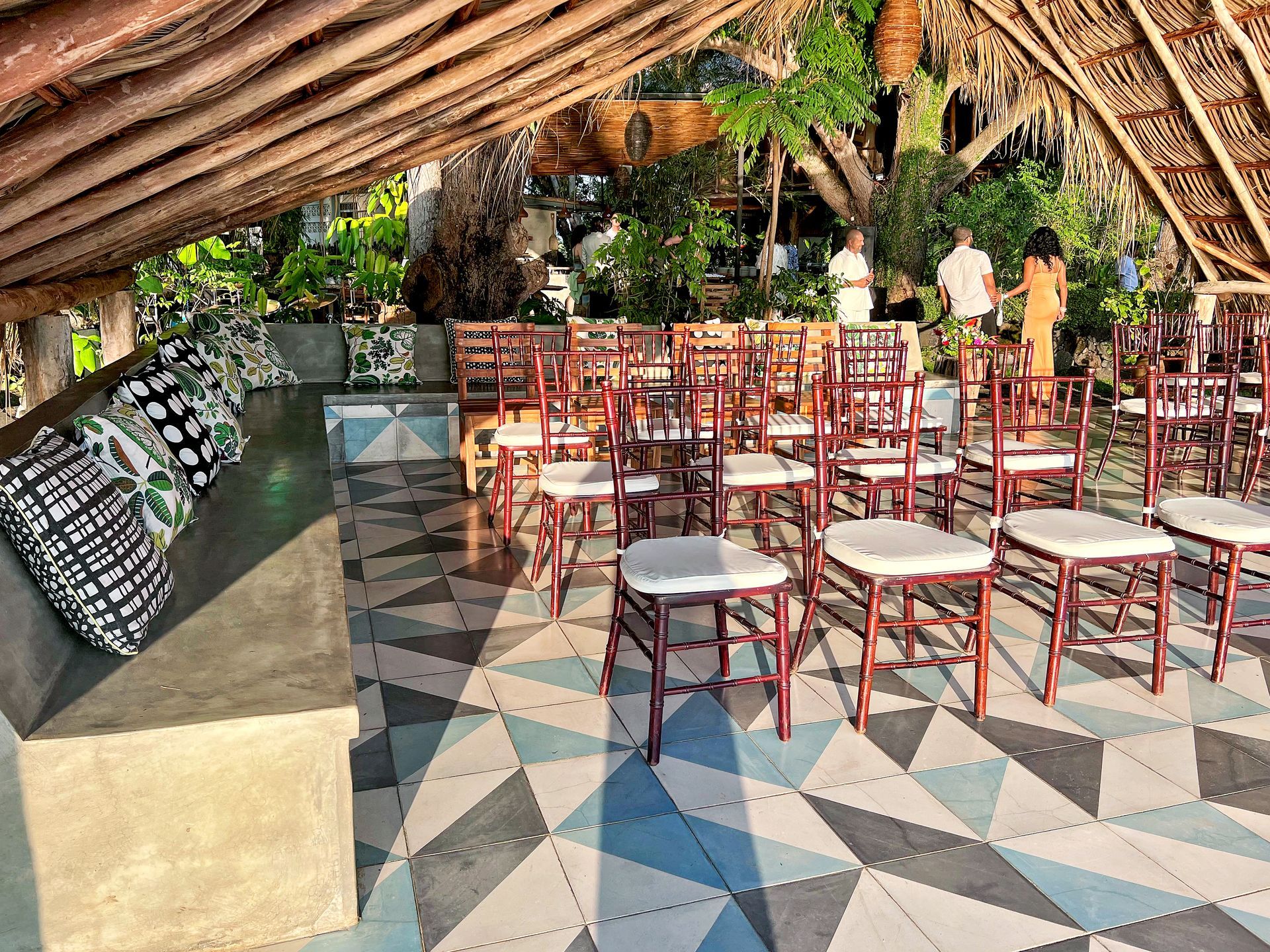 Ceremony seating under a thatched roof, geometric floor, with a couple and guests in the background.
