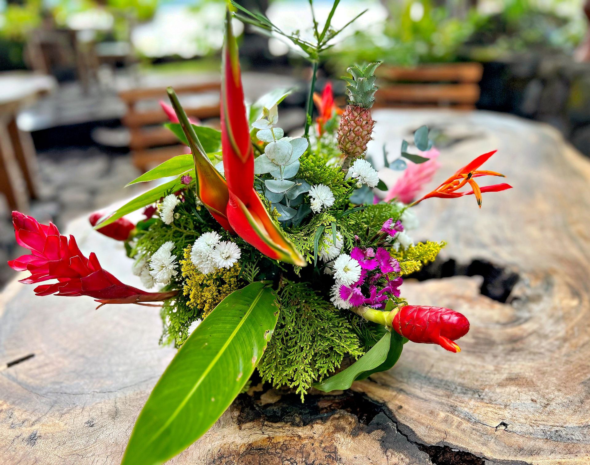 Tropical floral arrangement on a rustic wooden table, featuring red and green flowers with a small pineapple.