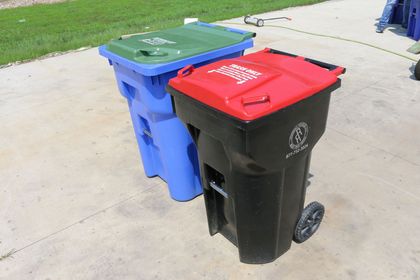 Three wheeled trash bins: blue, black, and a green lid bin. All are on a concrete surface outdoors.