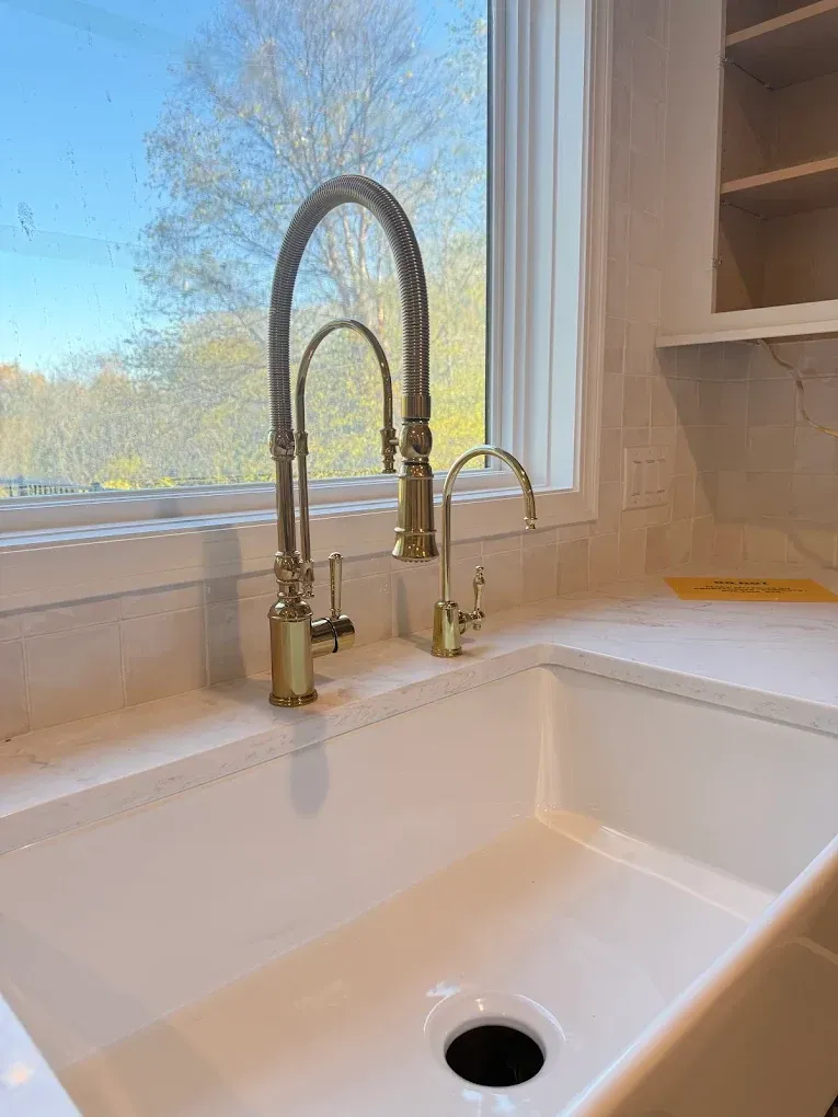Gold kitchen faucet over a white farmhouse sink, beside a window with a view of trees.