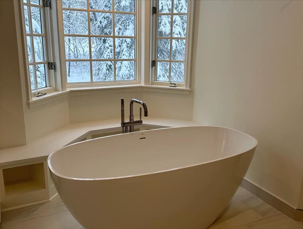 Freestanding white bathtub under a large window with trees visible. Modern bathroom with neutral walls.
