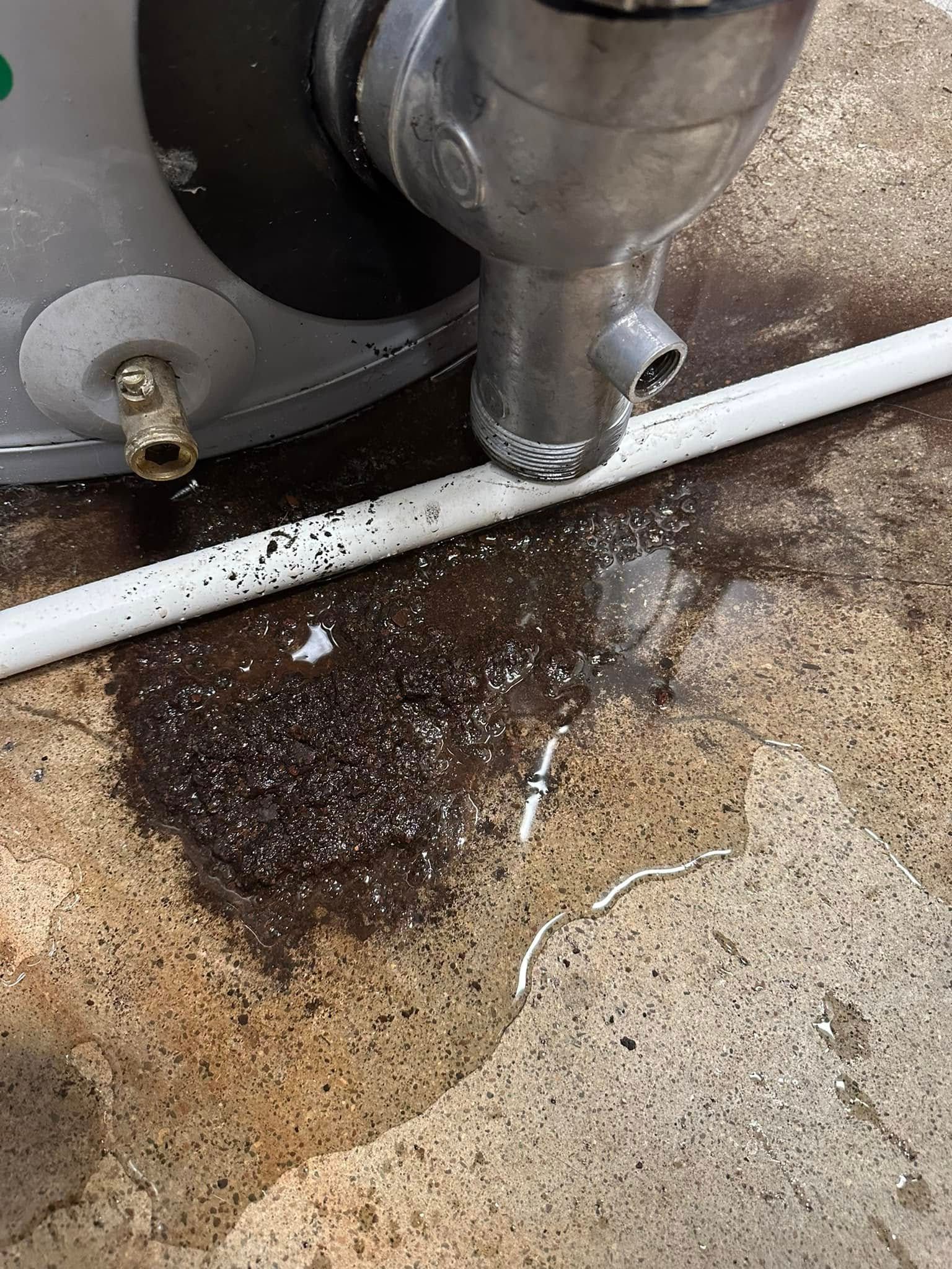 Leaking water heater with water pooling on concrete floor. White pipe nearby.
