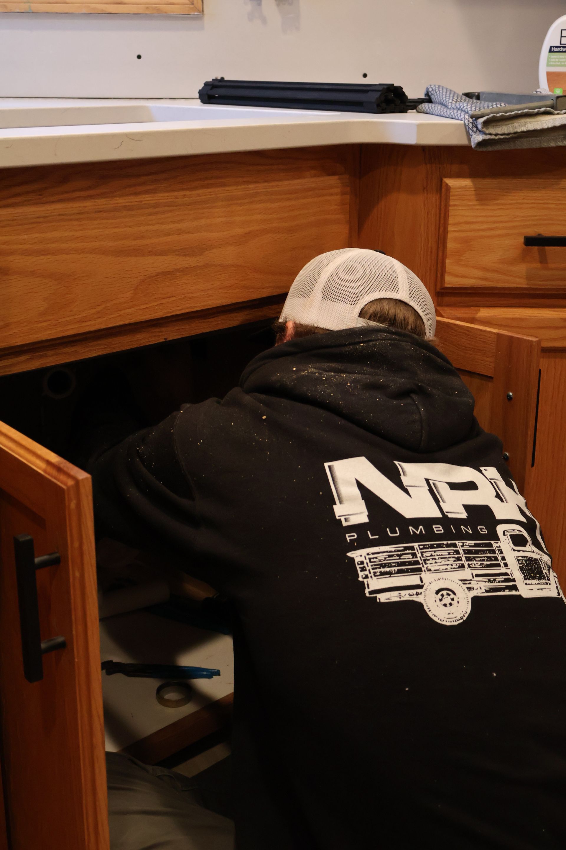 Two workers in orange and gray shirts install plumbing near a black water heater in a basement.