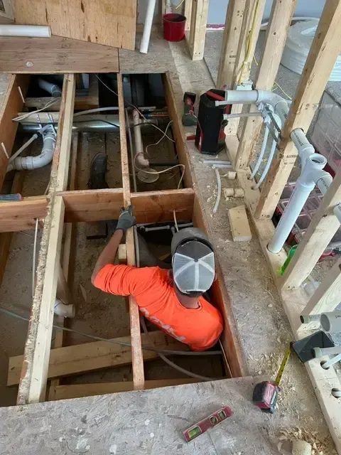 Construction worker installing plumbing pipes in a floor. Interior framing, orange shirt, and safety mask visible.