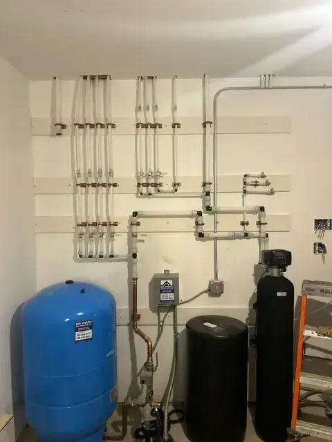 Water pipes and tanks mounted on a white wall. Includes a blue tank, black tanks, and copper/plastic piping.