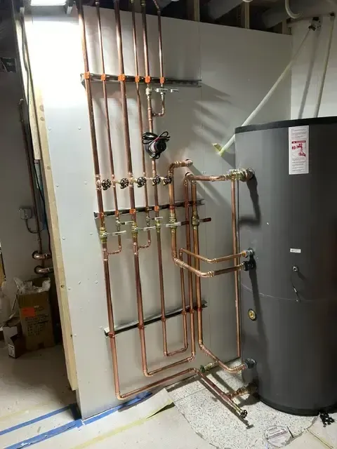 Copper pipes and fittings installed on a white wall next to a large black water heater tank.