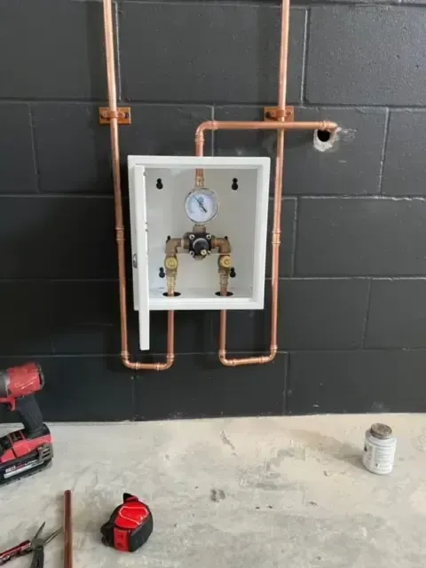 Copper pipes and valve assembly in white box on black tiled wall during construction.
