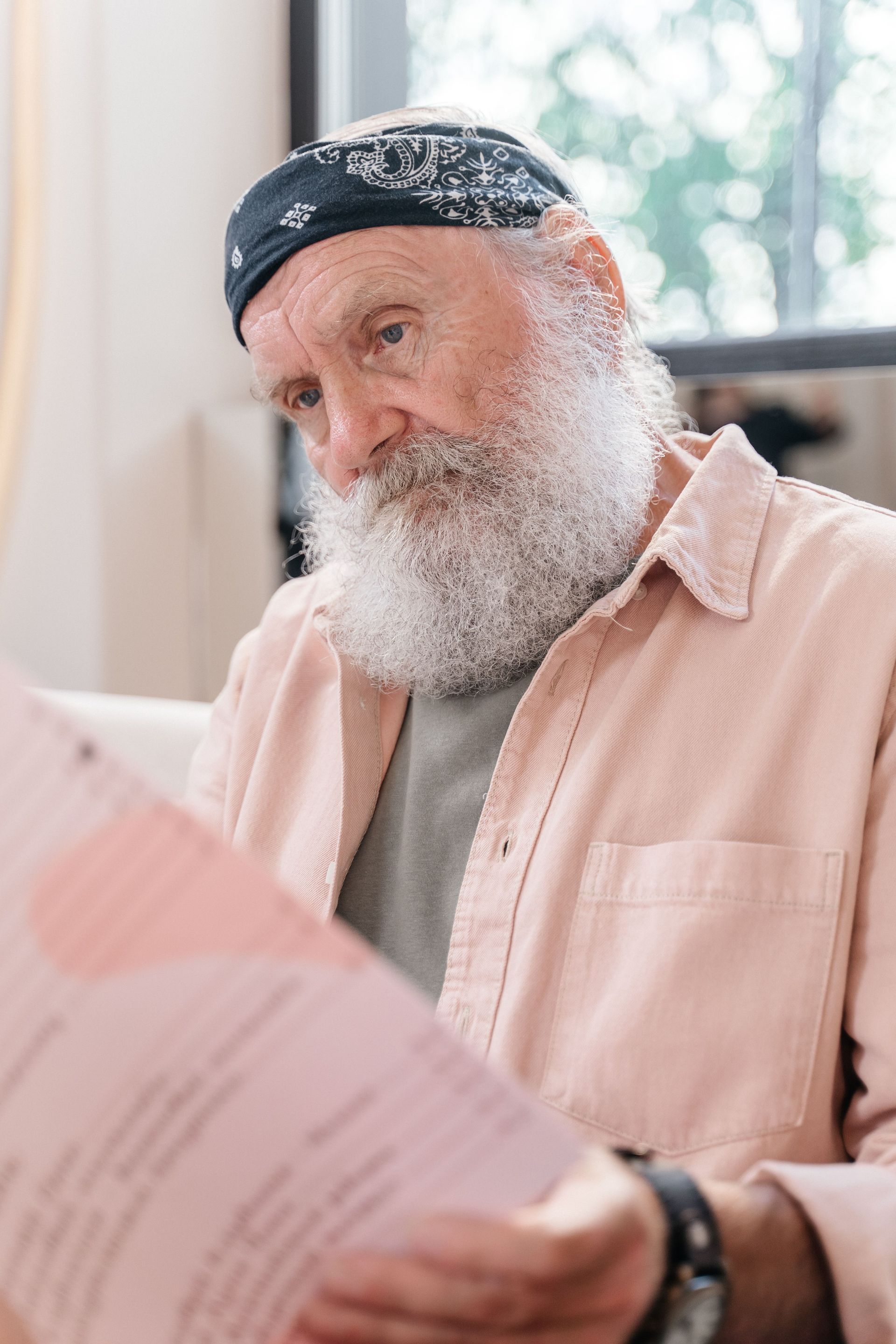Bearded person in a bandana reading papers by a window in a bright room