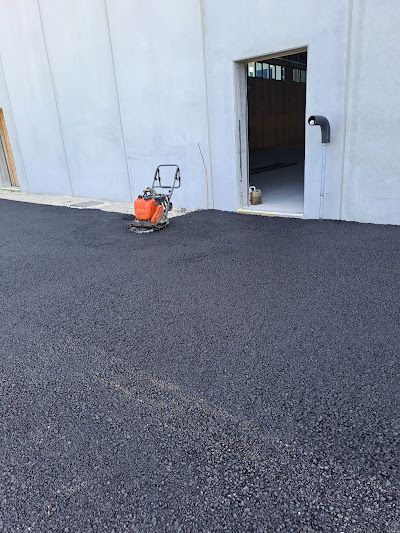 Una piastra vibrante arancione è posizionata su una superficie di asfalto appena posata, all'esterno dell'ingresso di un edificio.