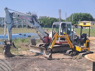 Un grande escavatore bianco e un escavatore JCB più piccolo di colore giallo parcheggiati in un cantiere sterrato.