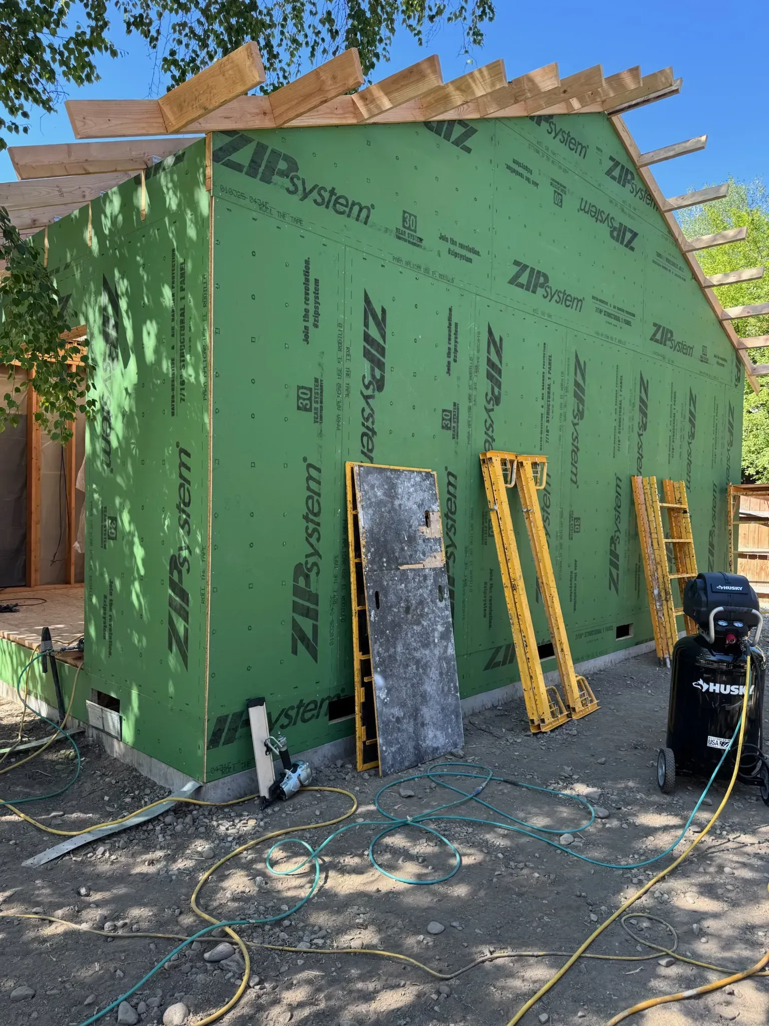 Green house under construction with exposed roof framing, ladders leaning outside, and tools on the ground.