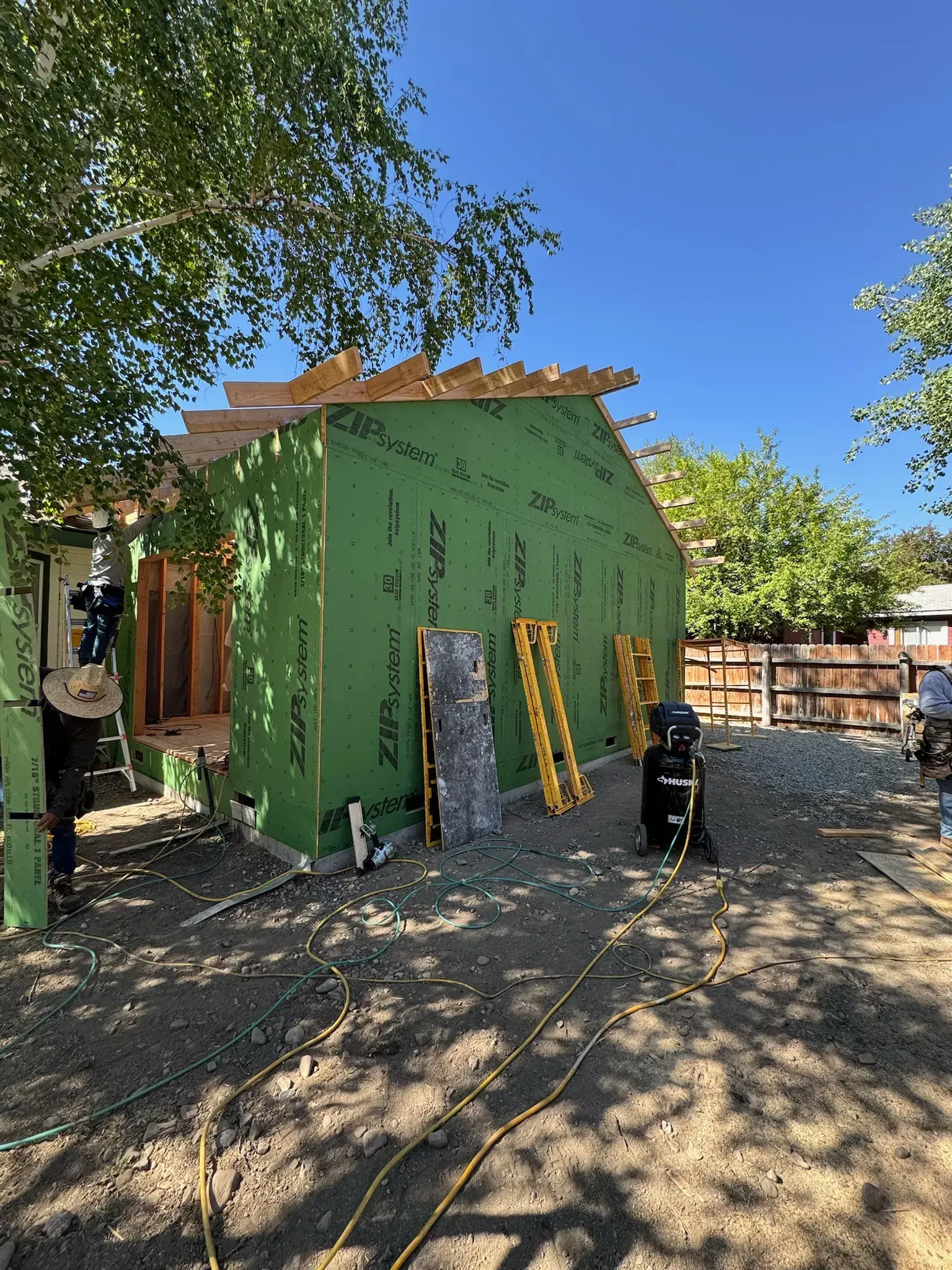 Green house under construction with exposed wrap, leaning boards, and trees in a sunny yard