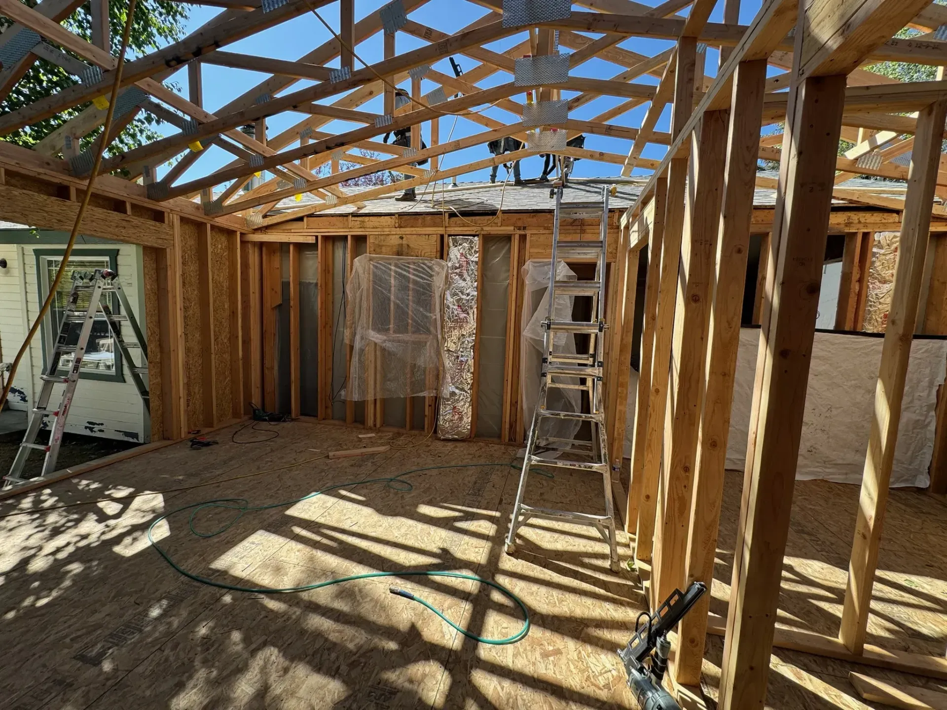 Interior of a house under construction with exposed wooden framing, ladders, and sunlight through the roof trusses.