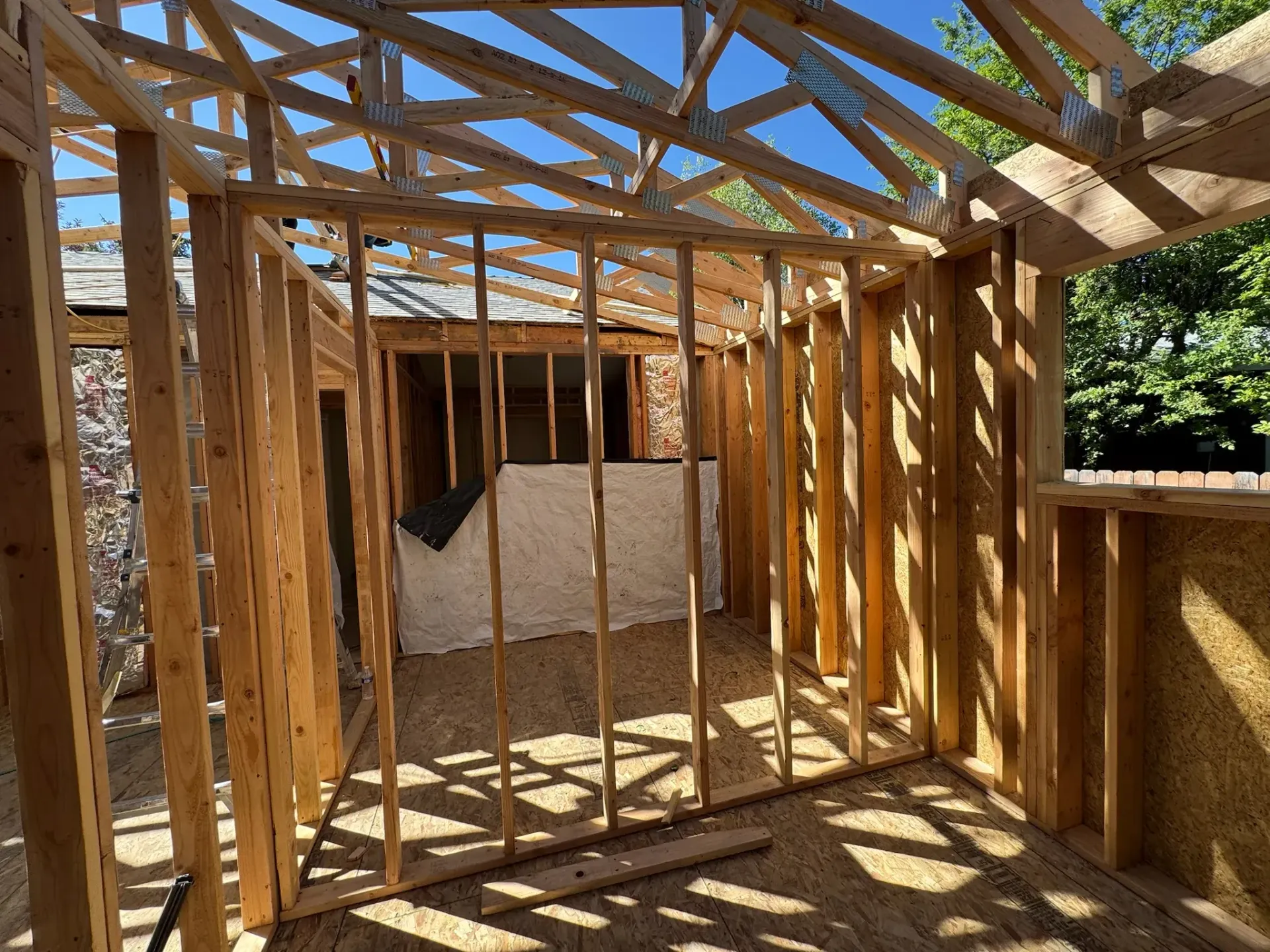 Interior of a wooden house under construction, with exposed framing and sunlight through the roof beams
