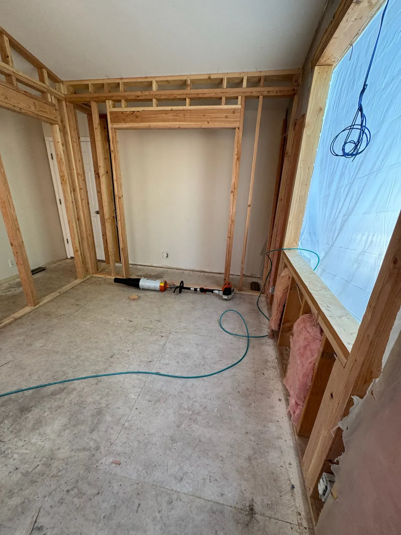 Framed room under construction with exposed studs, concrete floor, and a large window covered in plastic.