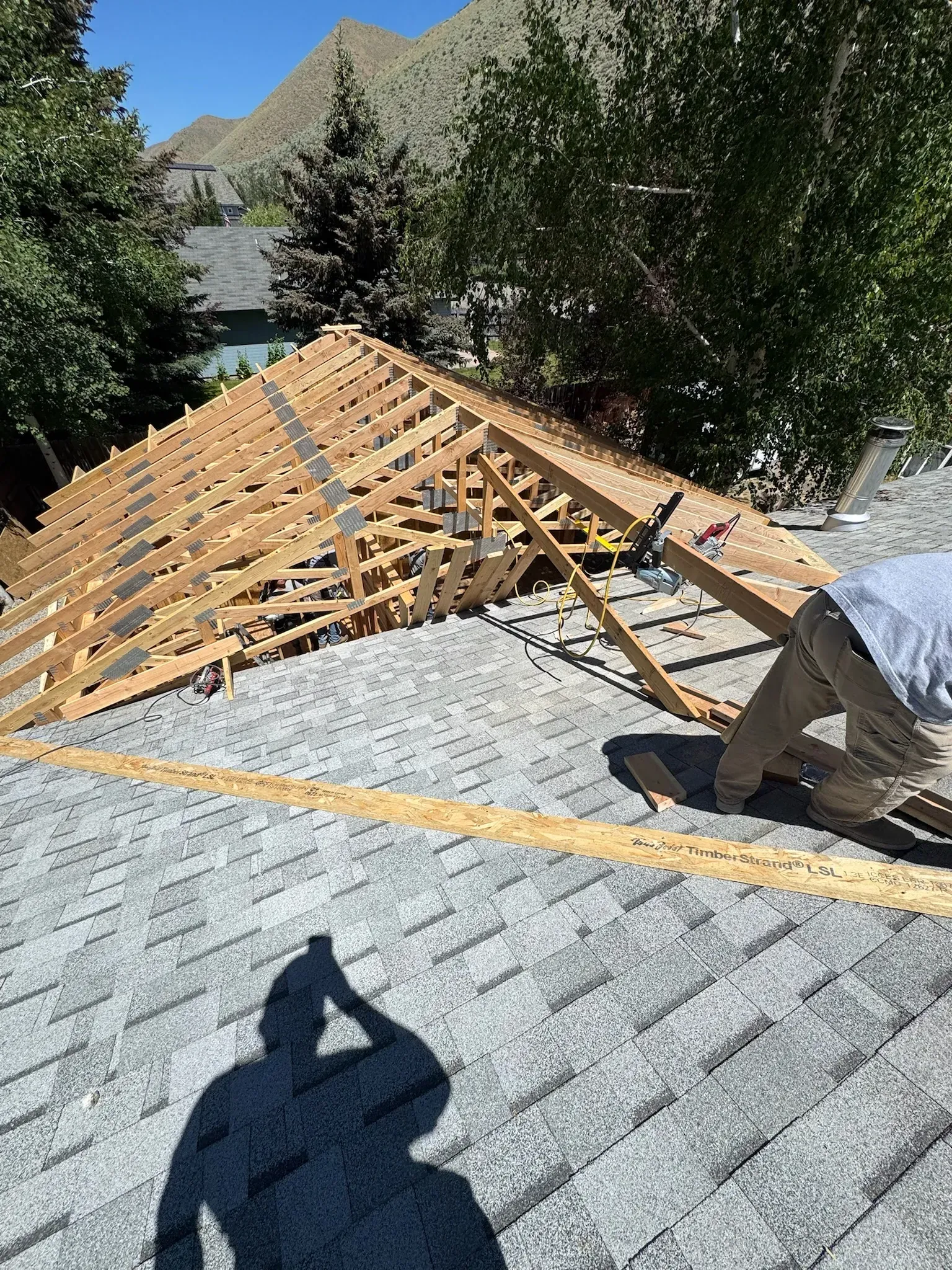Roof under construction with wooden trusses on a shingled house, viewed from above outdoors.