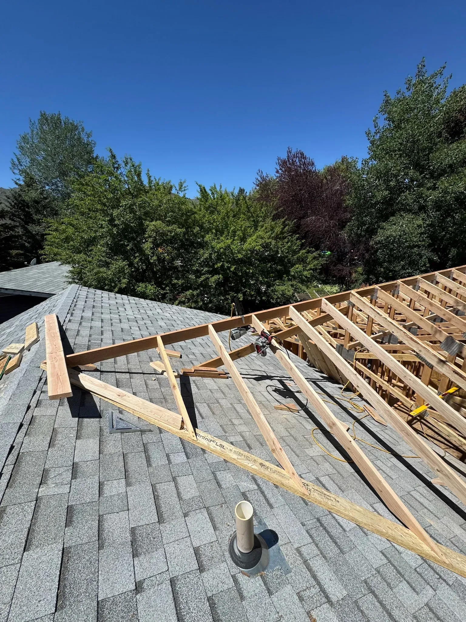 Rooftop with gray shingles and exposed wooden framing under a clear blue sky, trees in the background