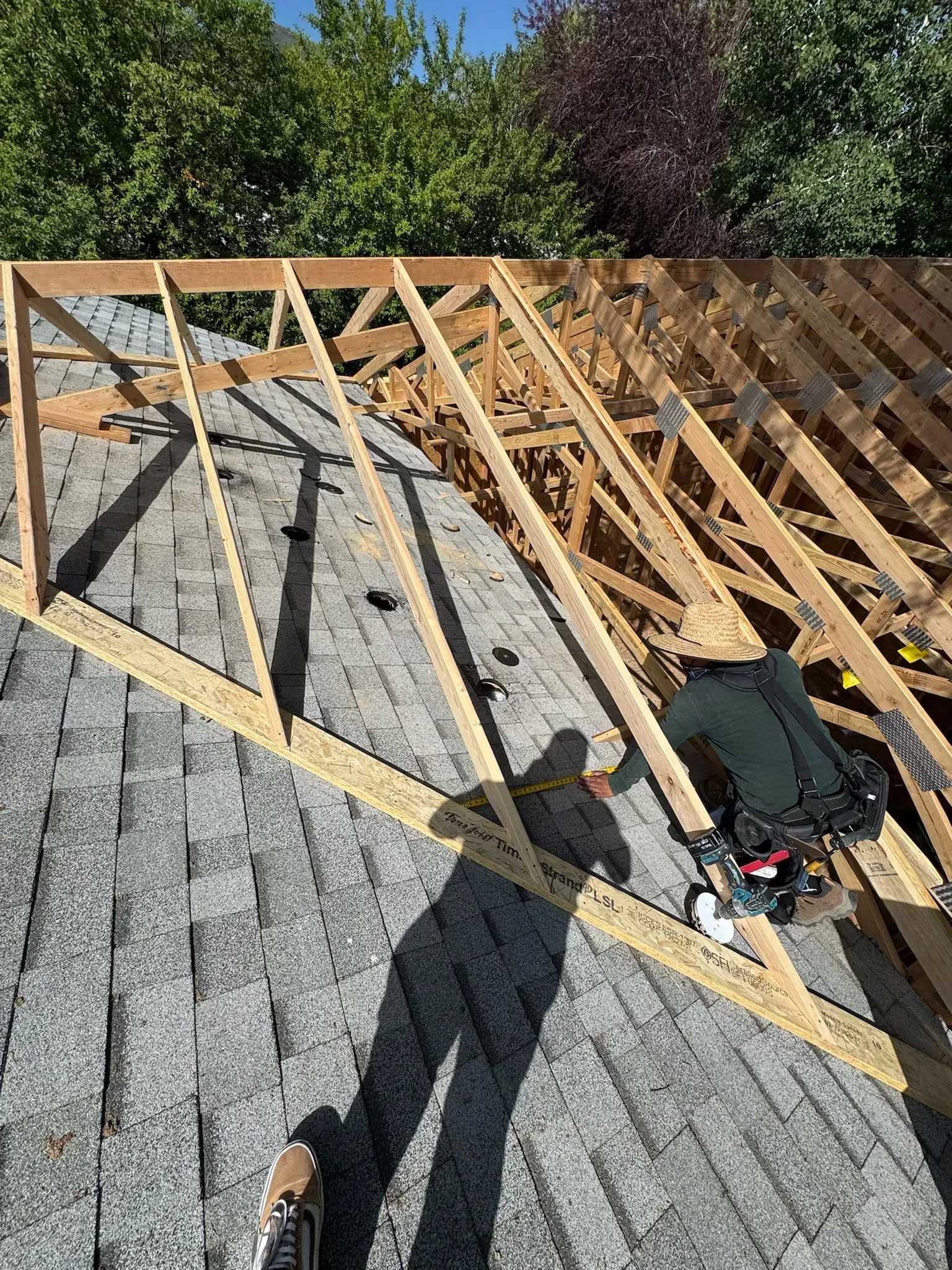 Roofing work in progress with exposed wooden trusses over gray shingles and a worker kneeling near the edge
