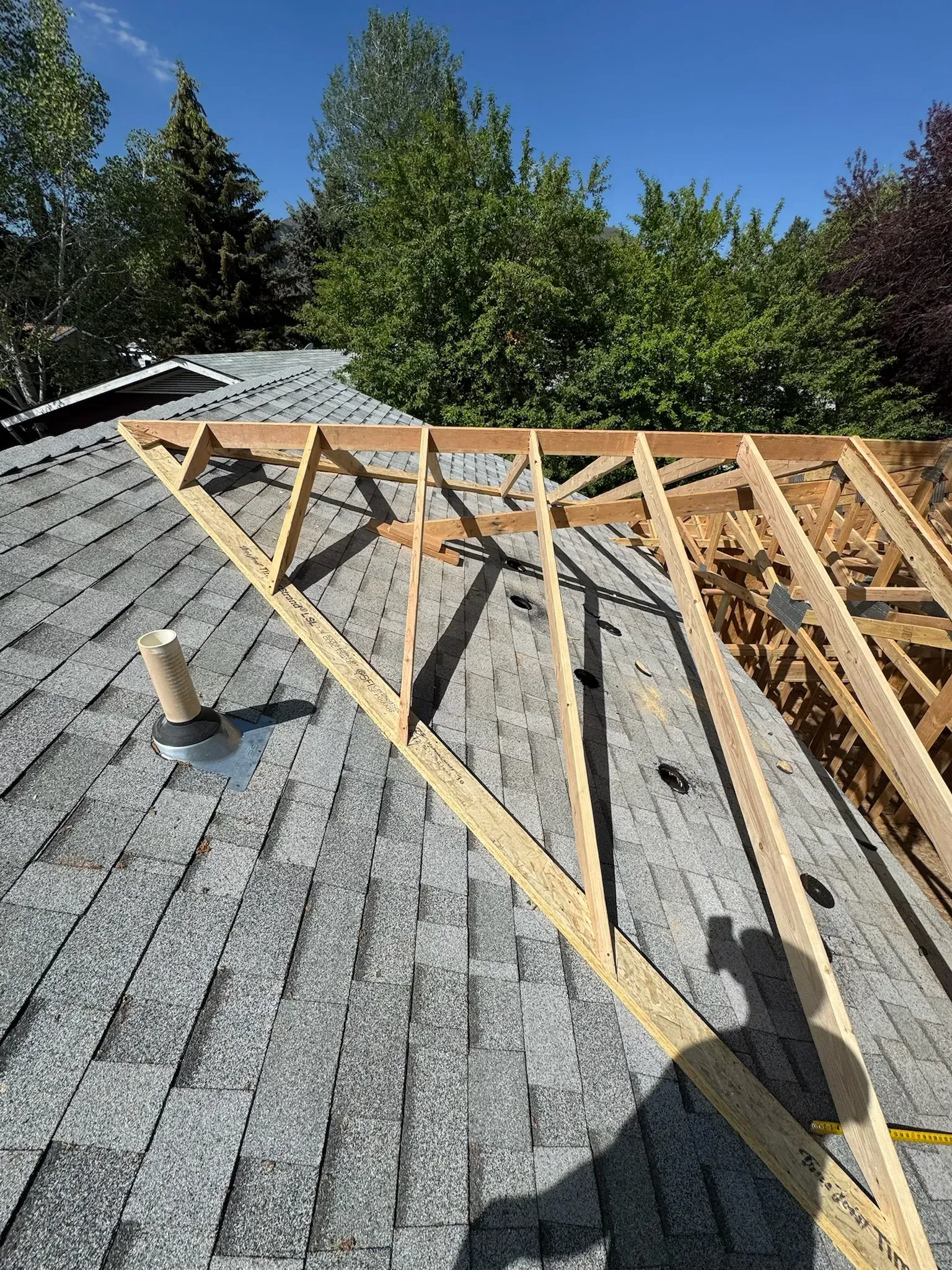 Roof framing under construction on a gray shingled roof, with wooden trusses and trees in the background
