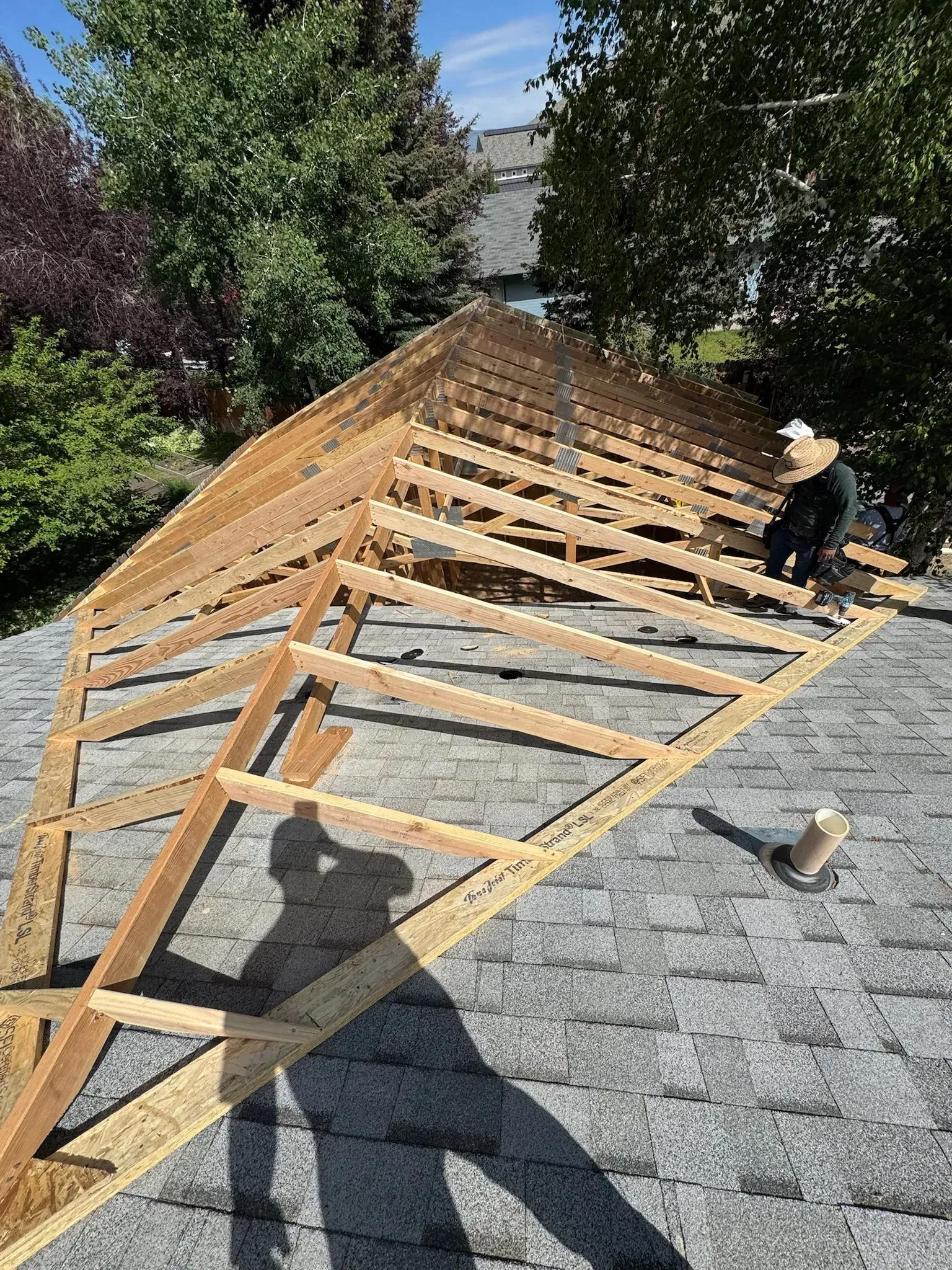 Person working on a roof with exposed wooden framing and gray shingles under blue sky.