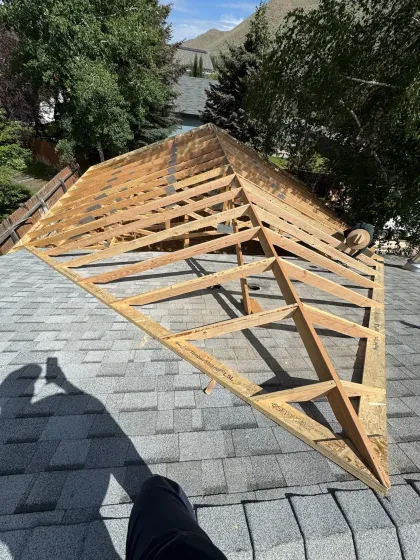 Roof framing underway on a shingled roof, with exposed wooden trusses beside trees and mountains.