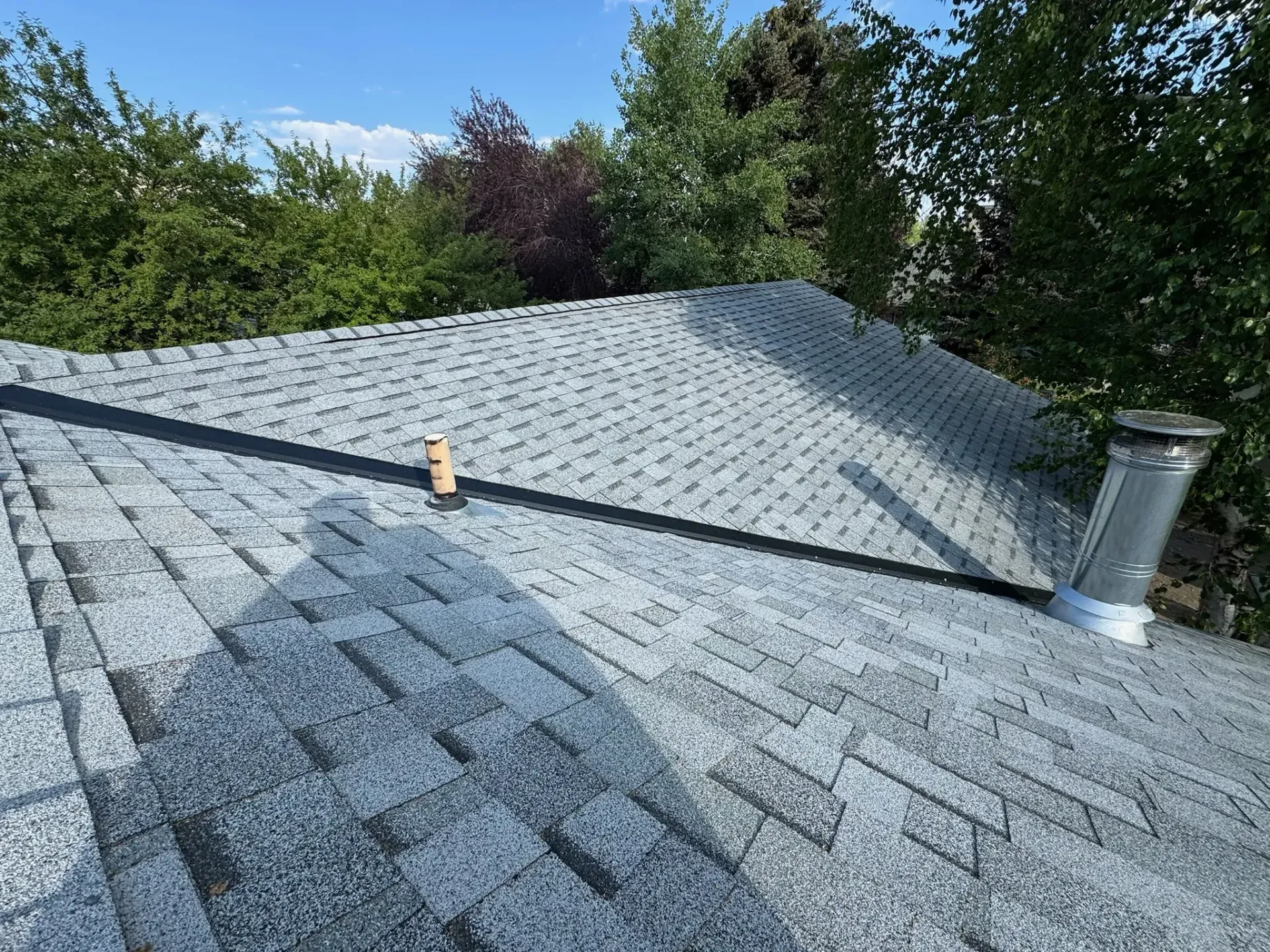 Gray shingled roof with a small vent pipe, surrounded by trees under a blue sky