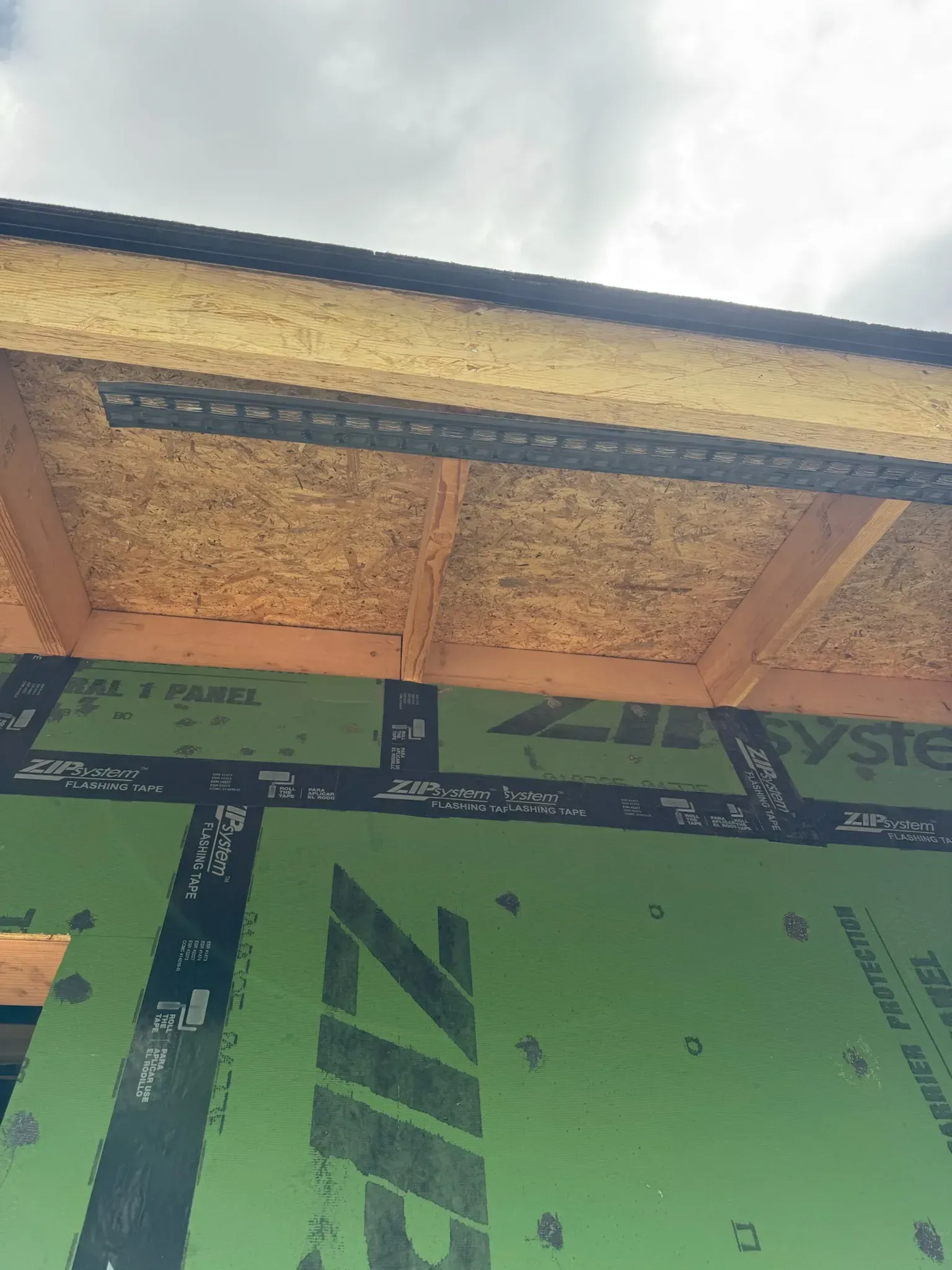 Partially built roof over green house wrap and exposed framing under a bright sky